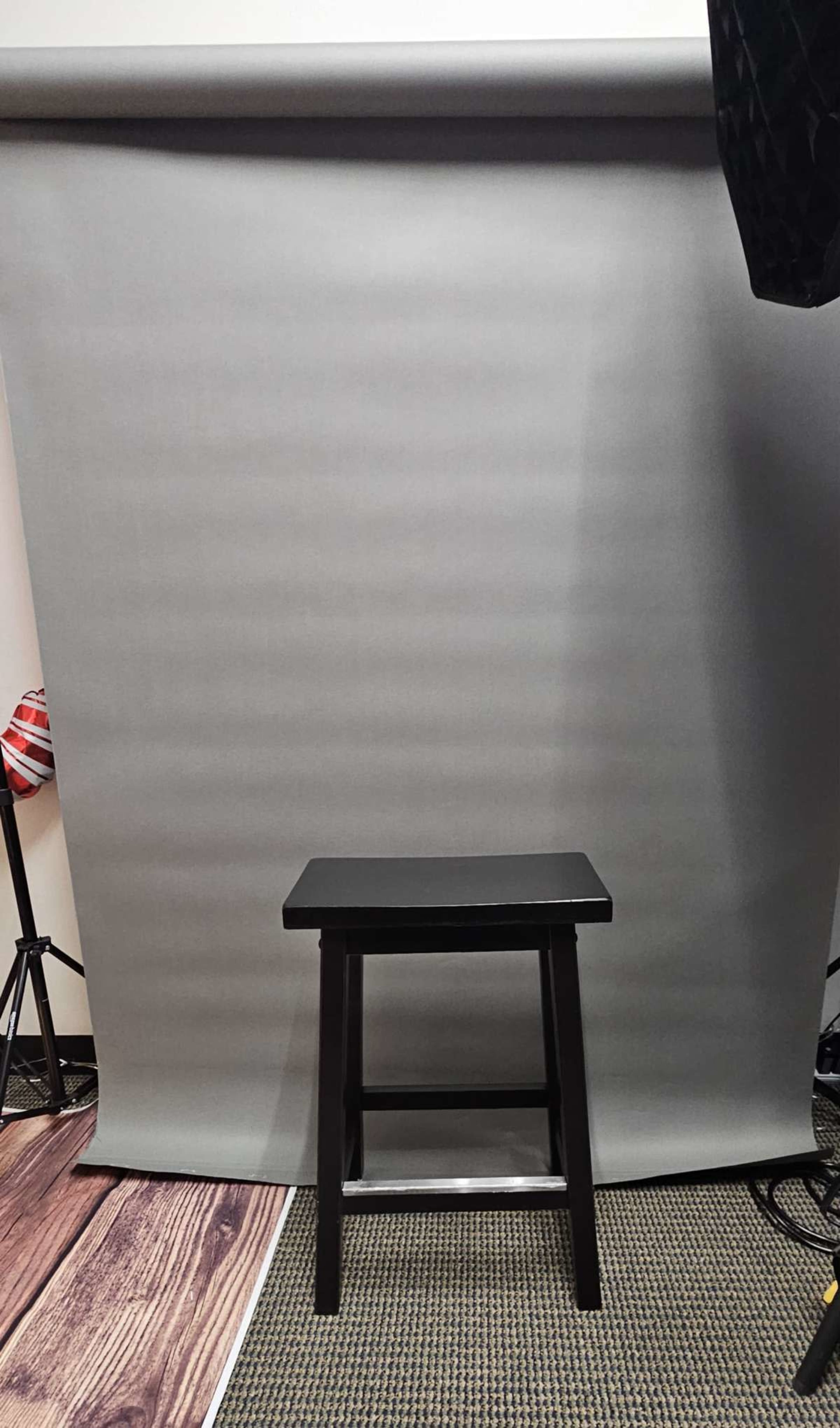 A black stool is positioned in front of a gray backdrop with studio lighting equipment nearby.