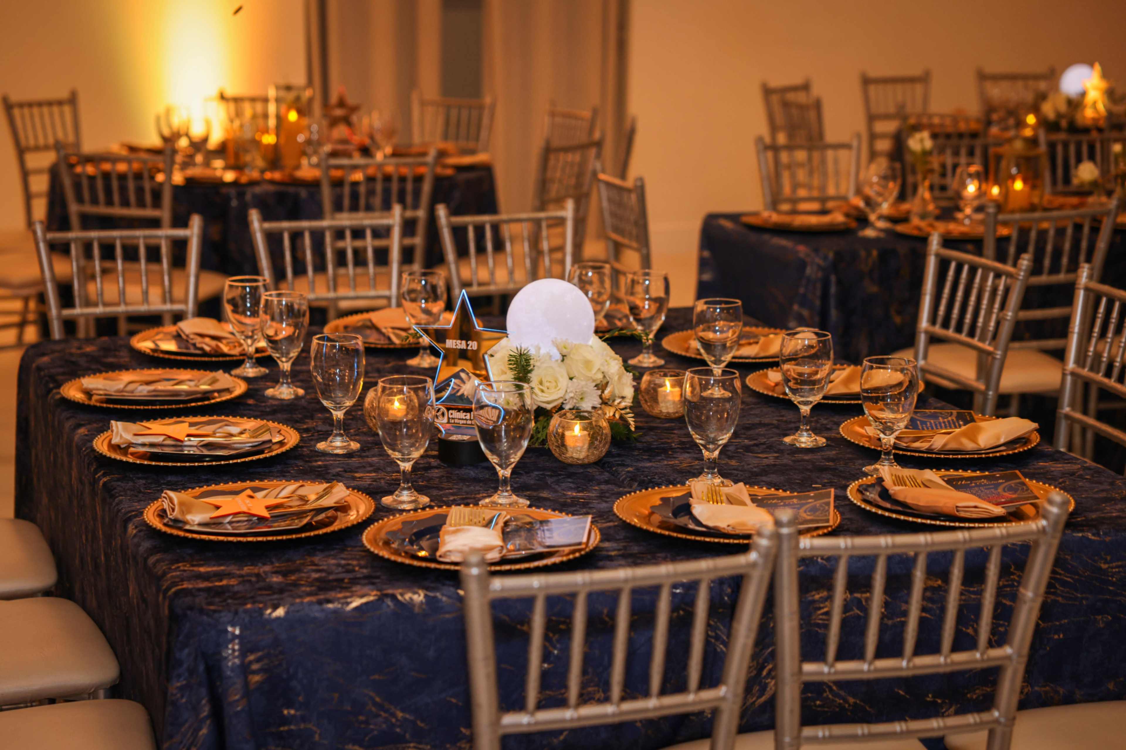 The image shows a formal dining setup with elegantly arranged tables featuring gold and navy tablecloths, crystal glassware, and decorative centerpieces.