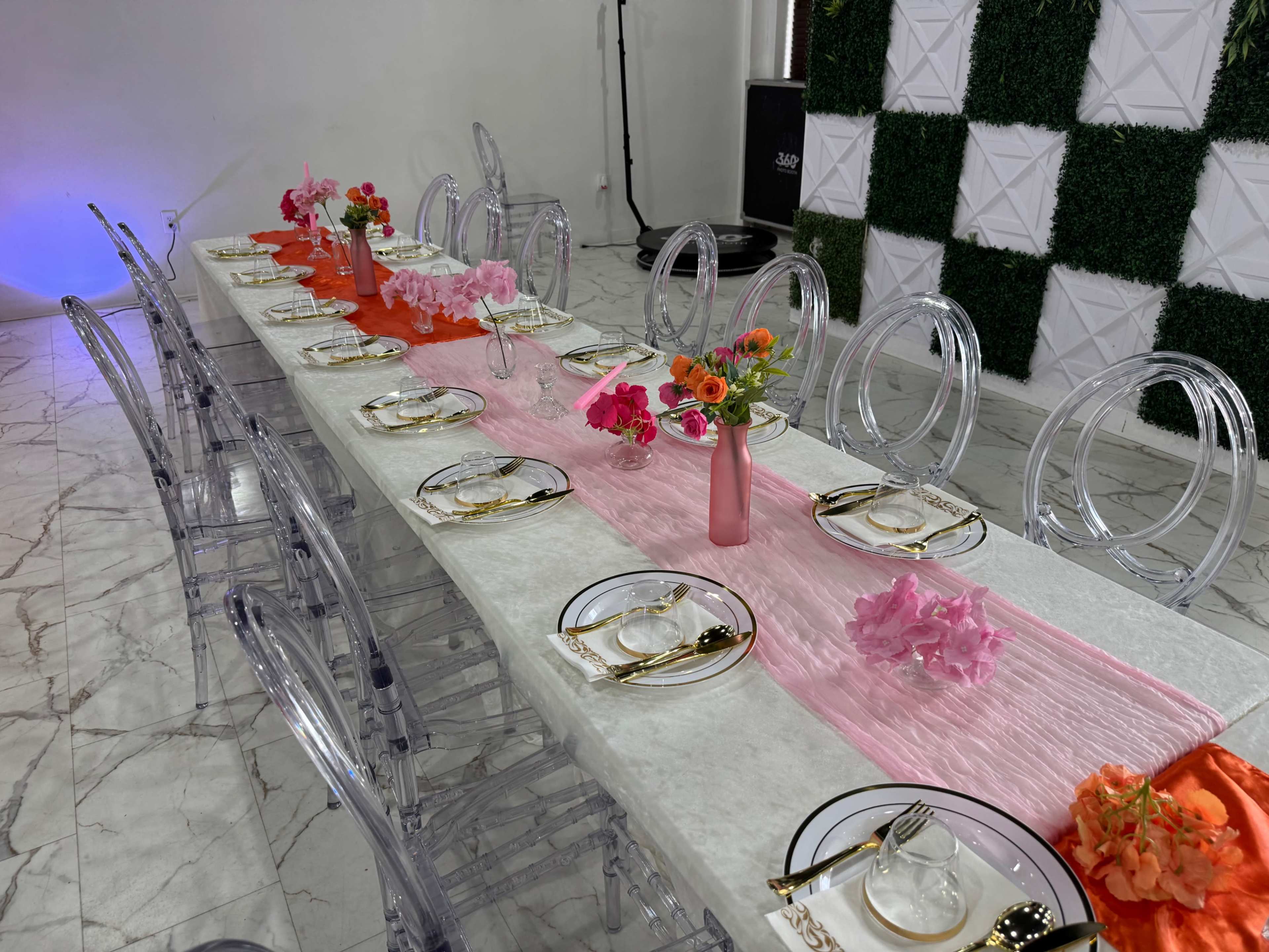 A long dining table is set with clear acrylic chairs, decorative runners in pink and orange, and floral centerpieces on a marble floor.