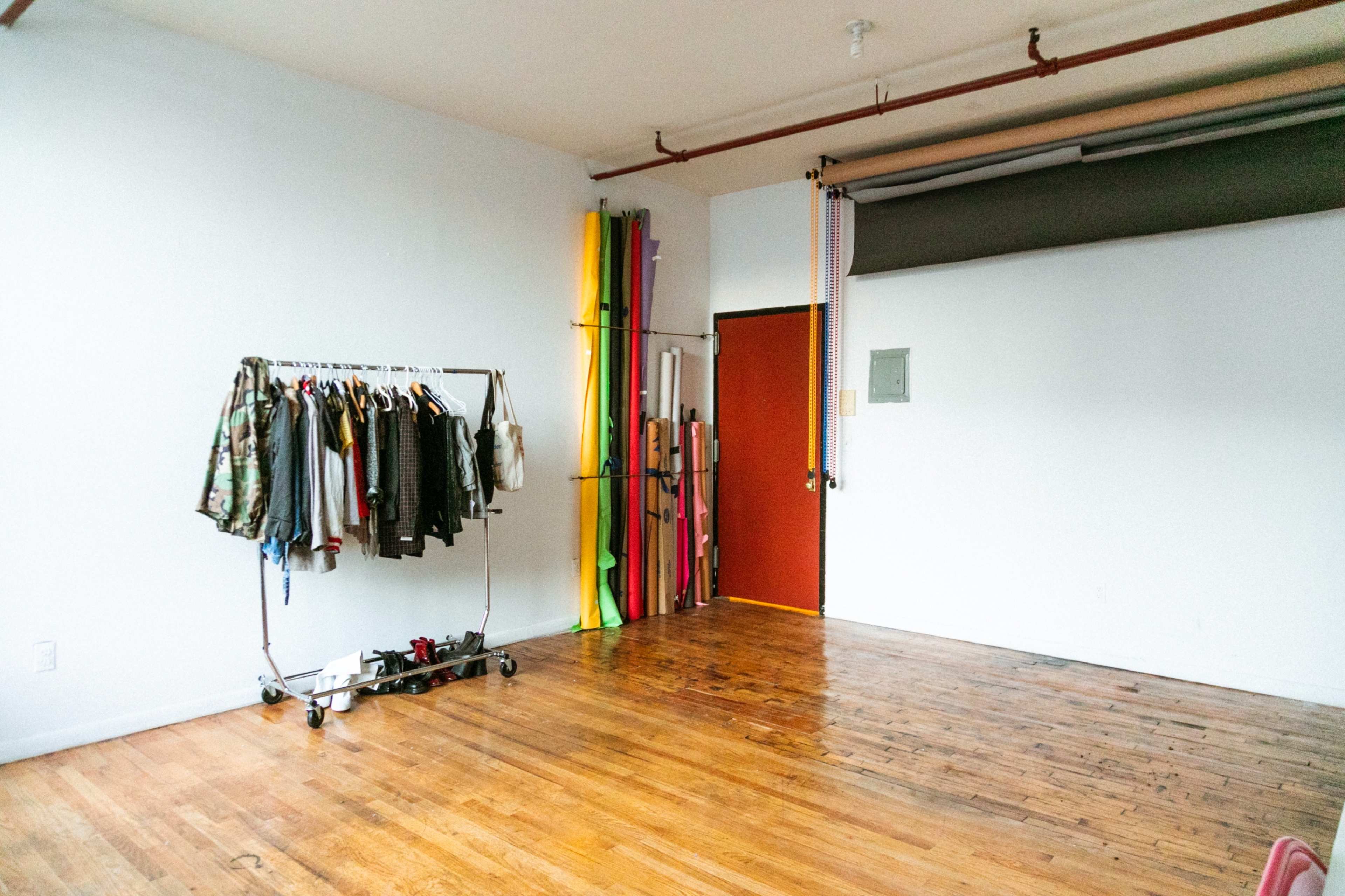 The image shows a spacious, empty room with wooden flooring, a clothing rack on one side, and colorful rolls of material leaning against a wall.