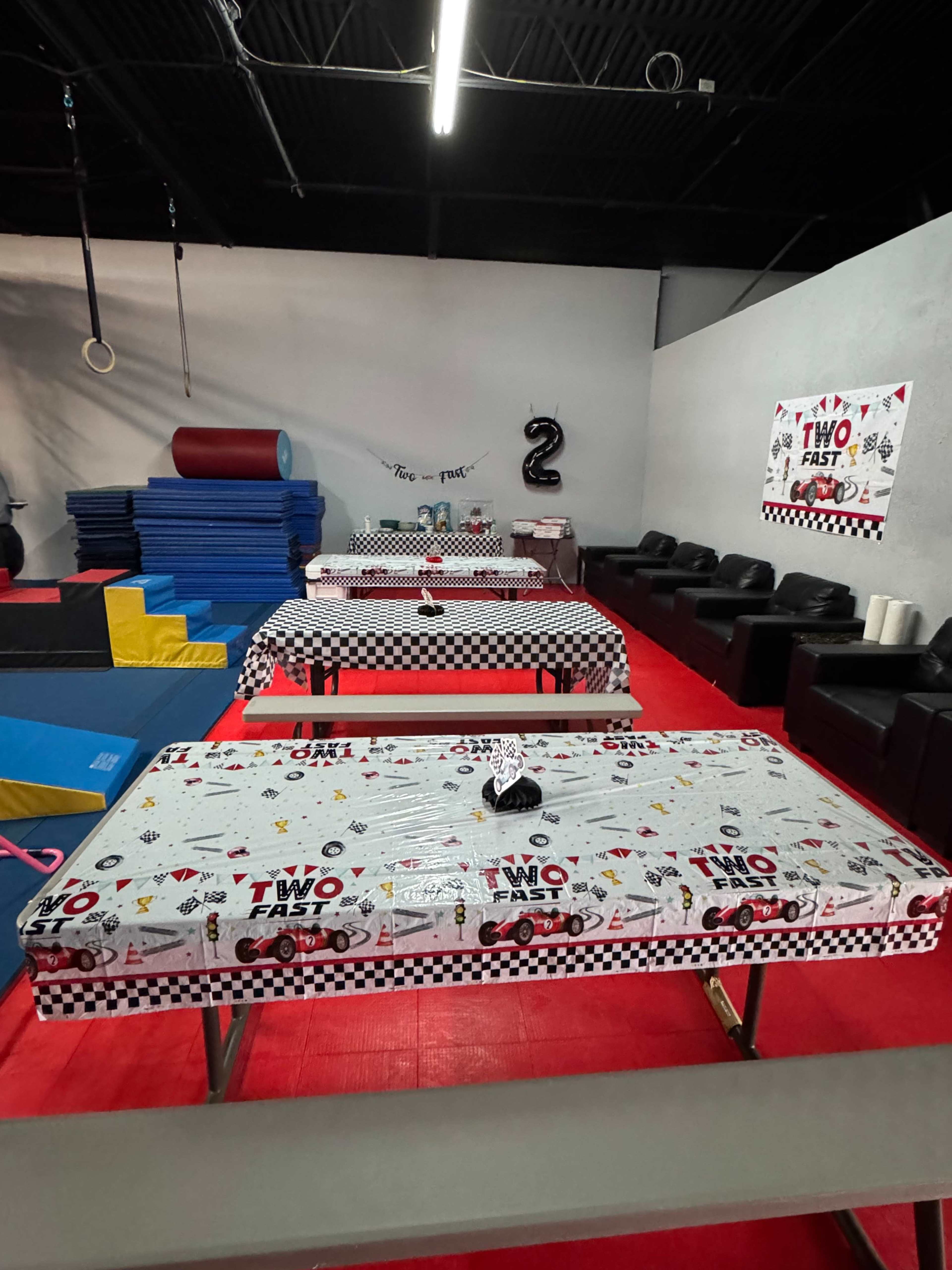 The image shows a colorful party setup with checkered tablecloths, decorated tables, and a wall featuring a "Two Fast" banner in a gym-like space.
