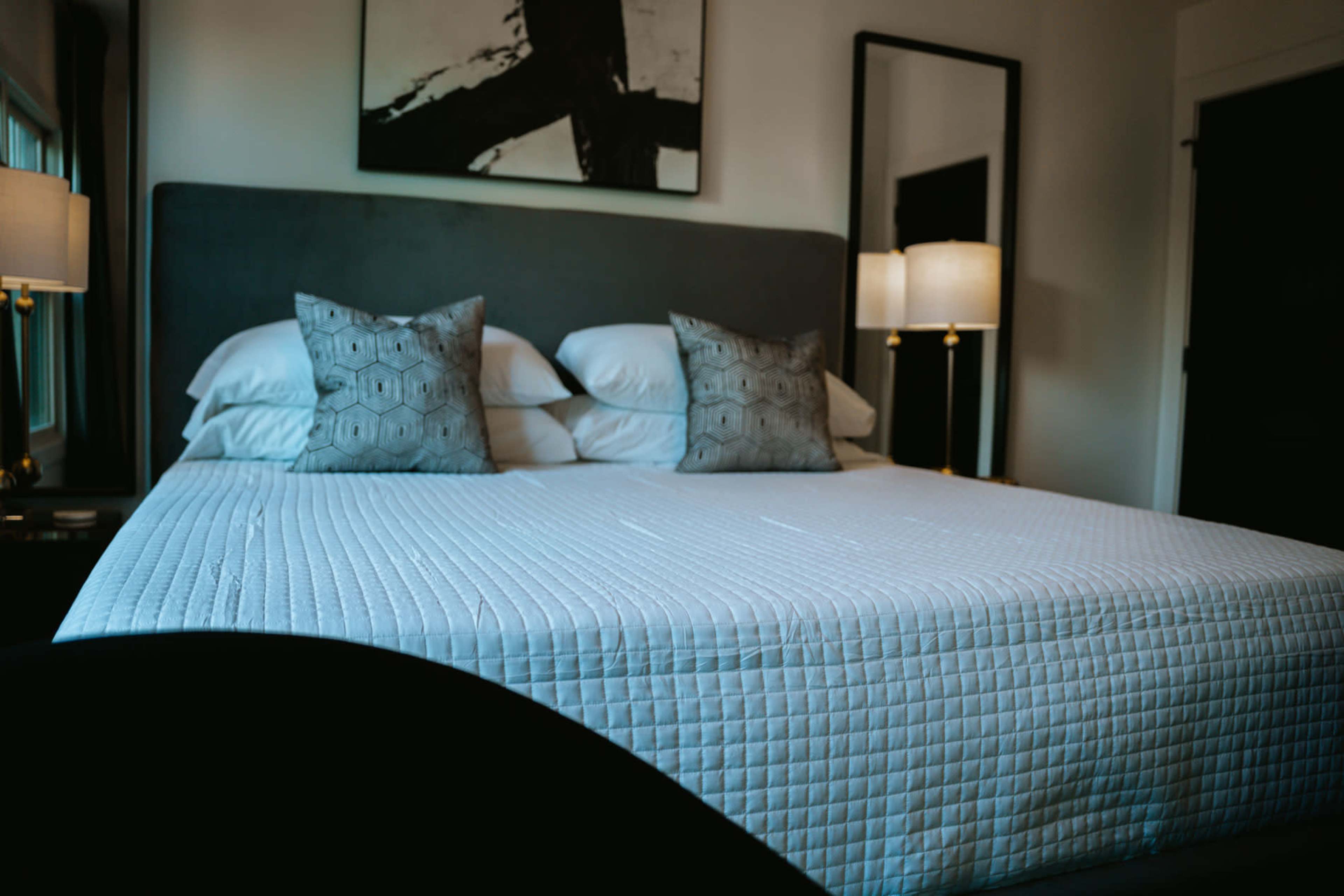 A neatly made bed with a light quilt, two decorative pillows, and a minimalistic bedroom setting featuring lamps and wall art.