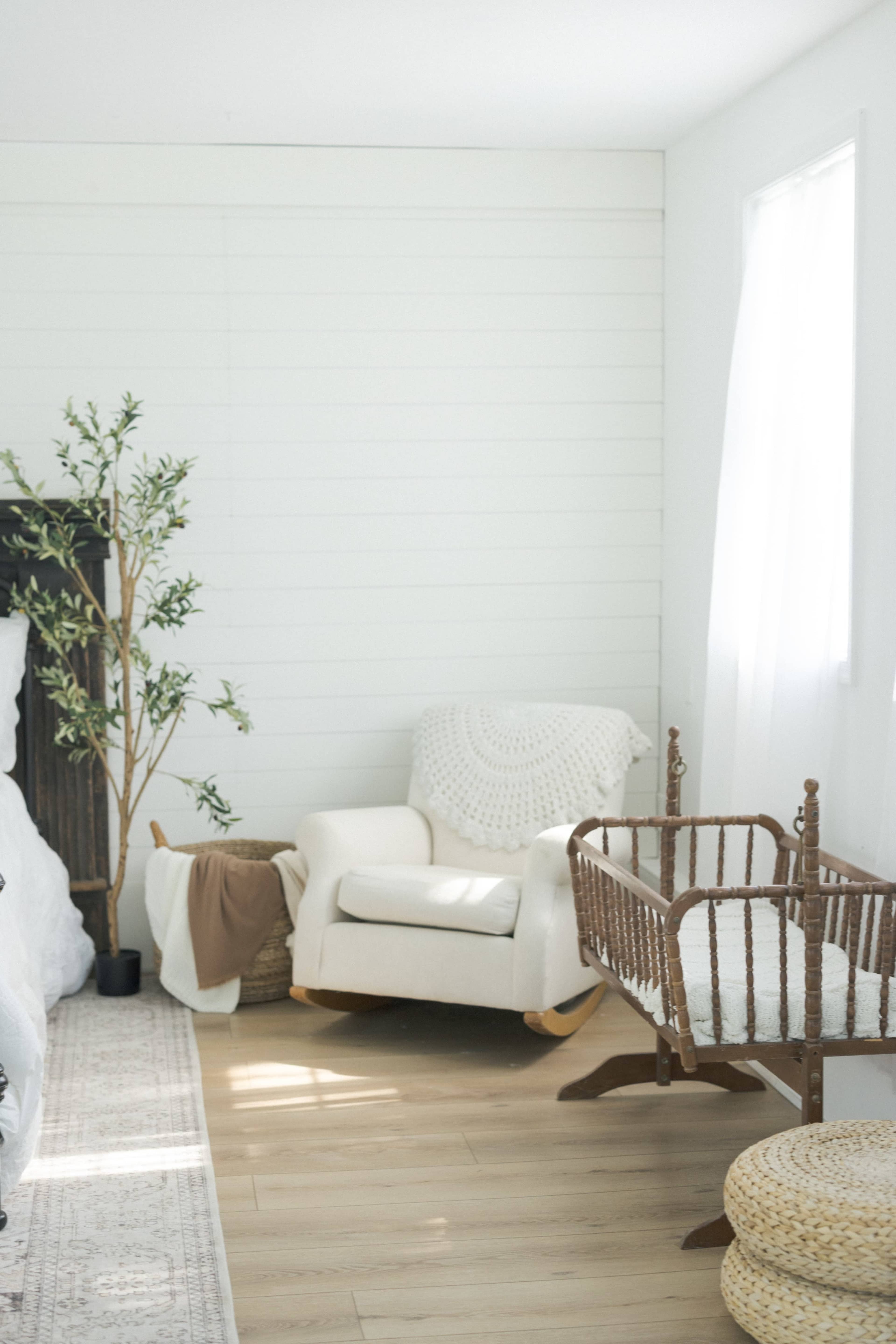 The image shows a bright, minimalistic bedroom with a white rocking chair, a woven baby crib, and a small plant beside them on a light wooden floor.