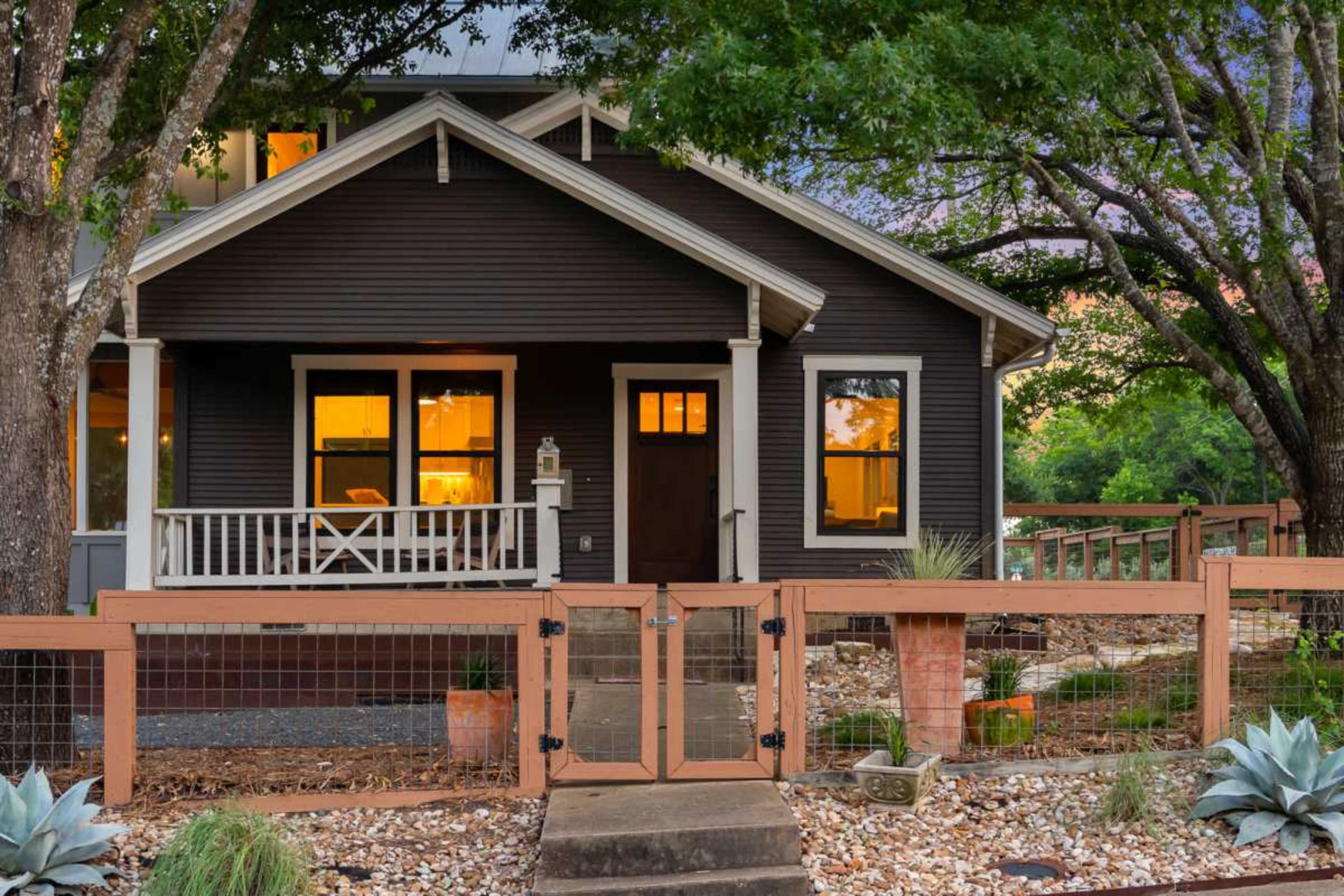 The image shows a dark-colored house with a porch, framed by trees, and fenced in a landscaped yard with gravel and potted plants.