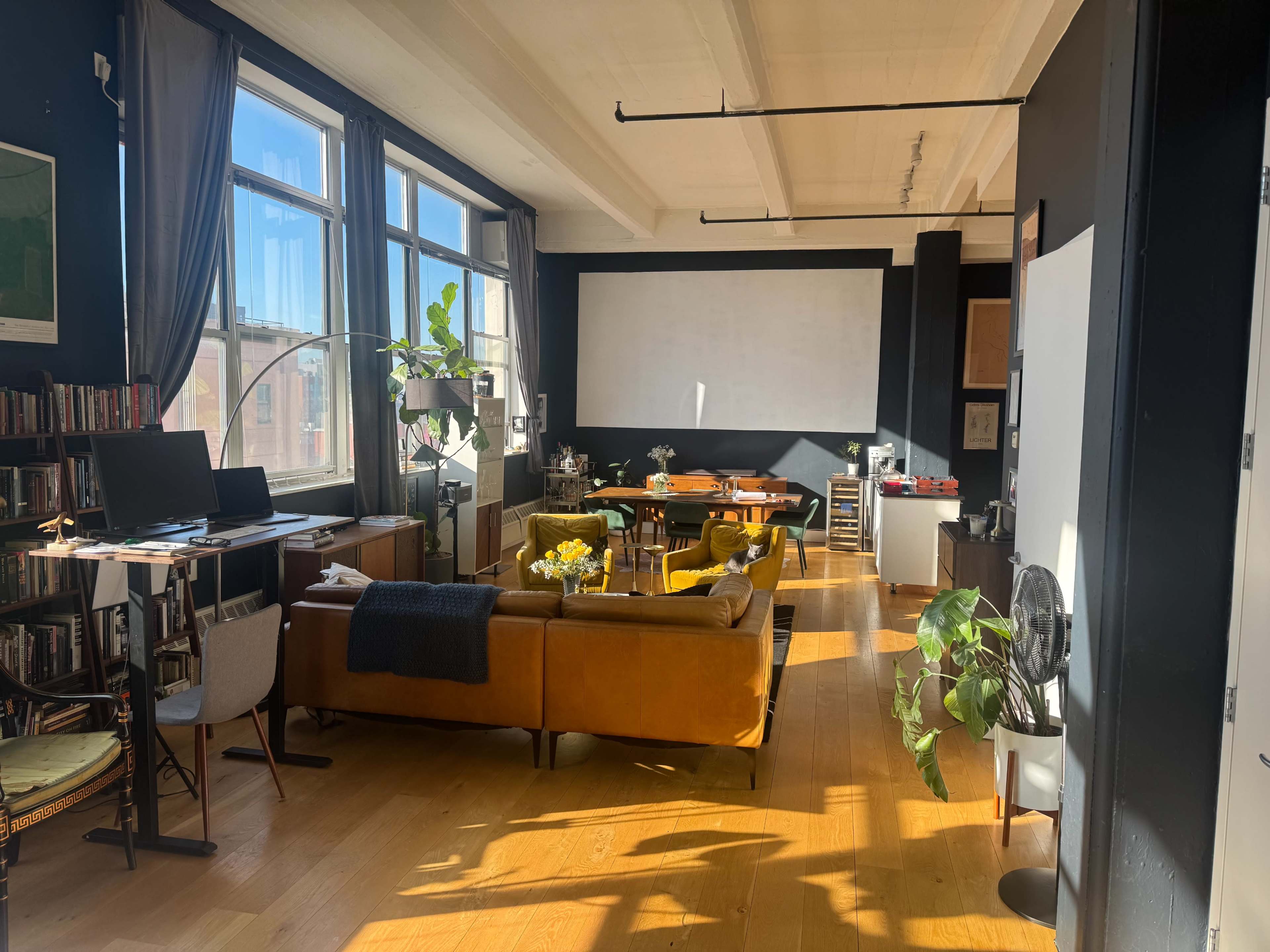 The image shows a spacious, well-lit apartment with large windows, a mix of modern and vintage furniture, and an open layout featuring a living area, workspace, and dining area.