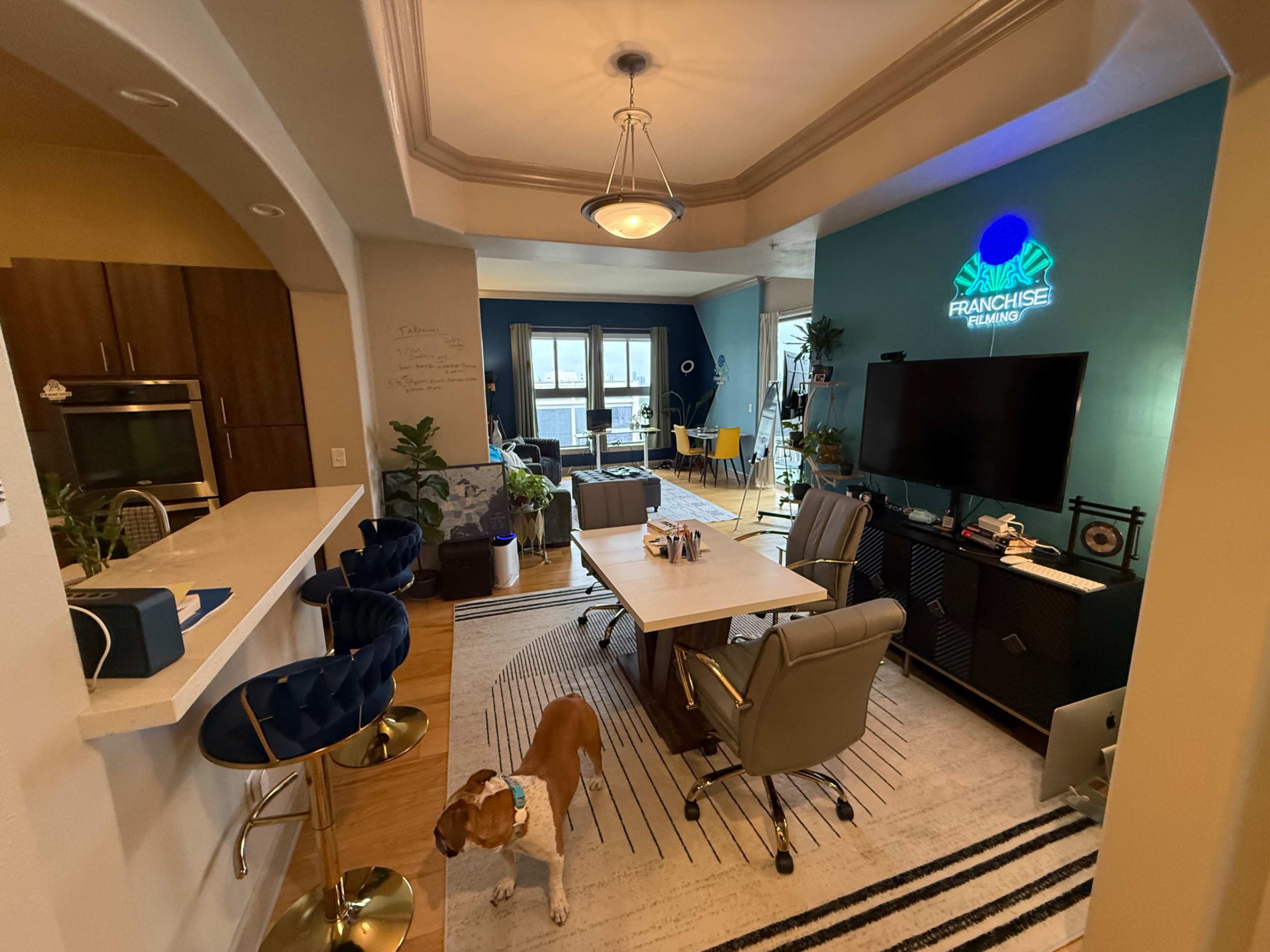 The image shows an open-concept modern office space with a dining area, including a large table, several chairs, and a dog on the floor.