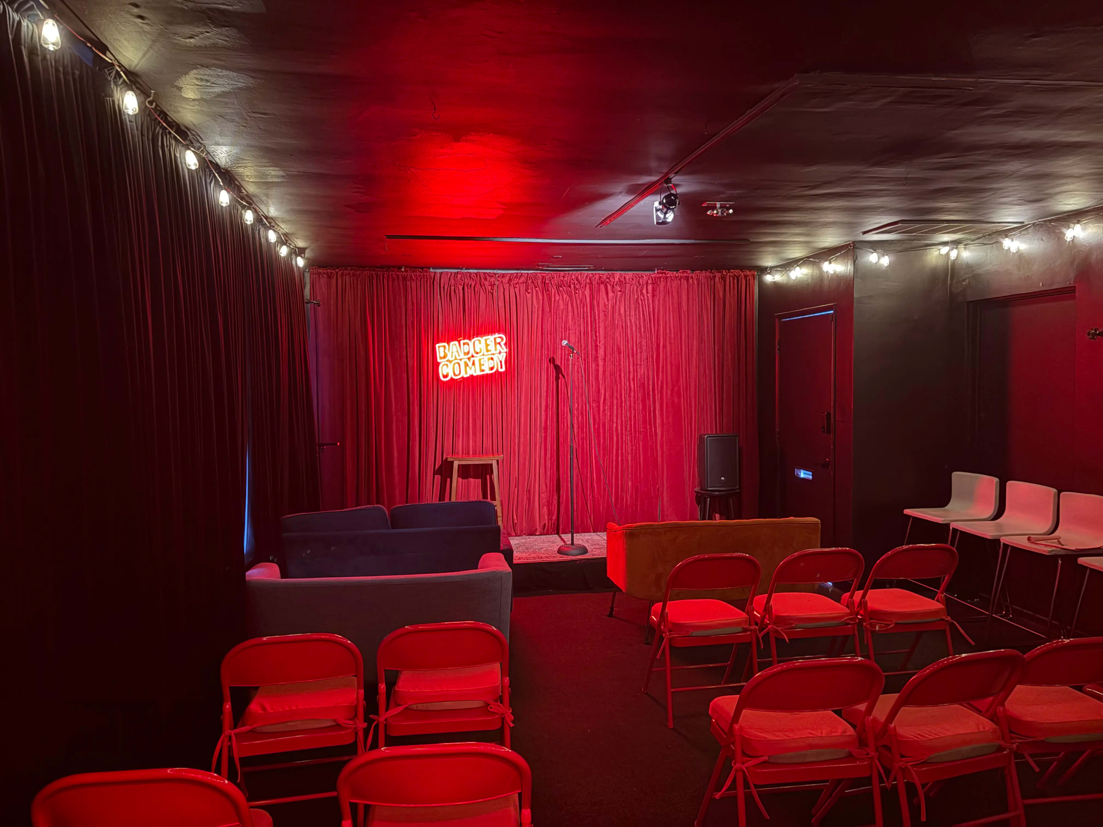 The image shows a small comedy venue with red walls, a stage featuring a microphone and stool, and rows of red chairs arranged for an audience.