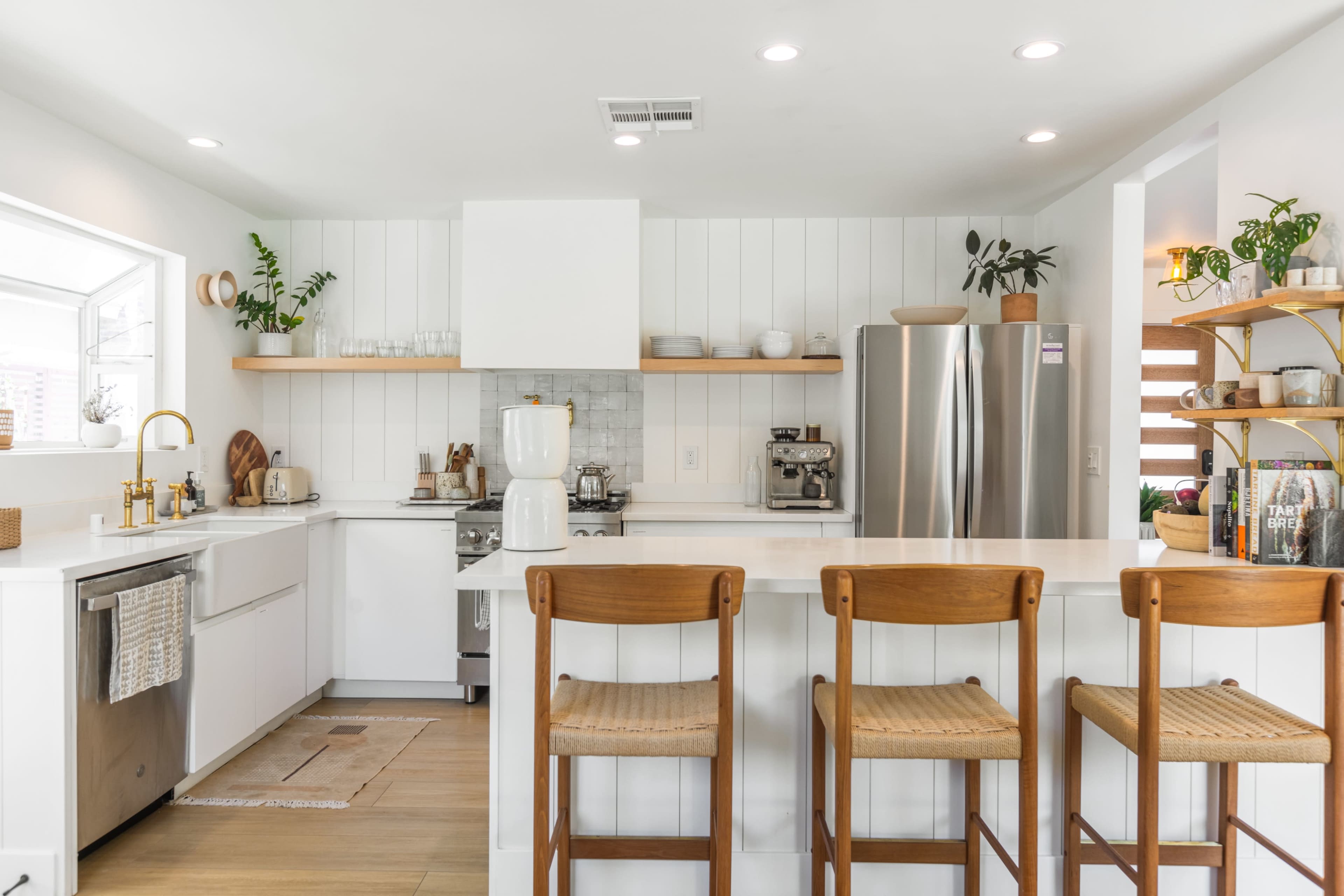 A modern kitchen features white cabinetry, a stainless steel refrigerator, and wooden bar stools arranged at a breakfast counter.