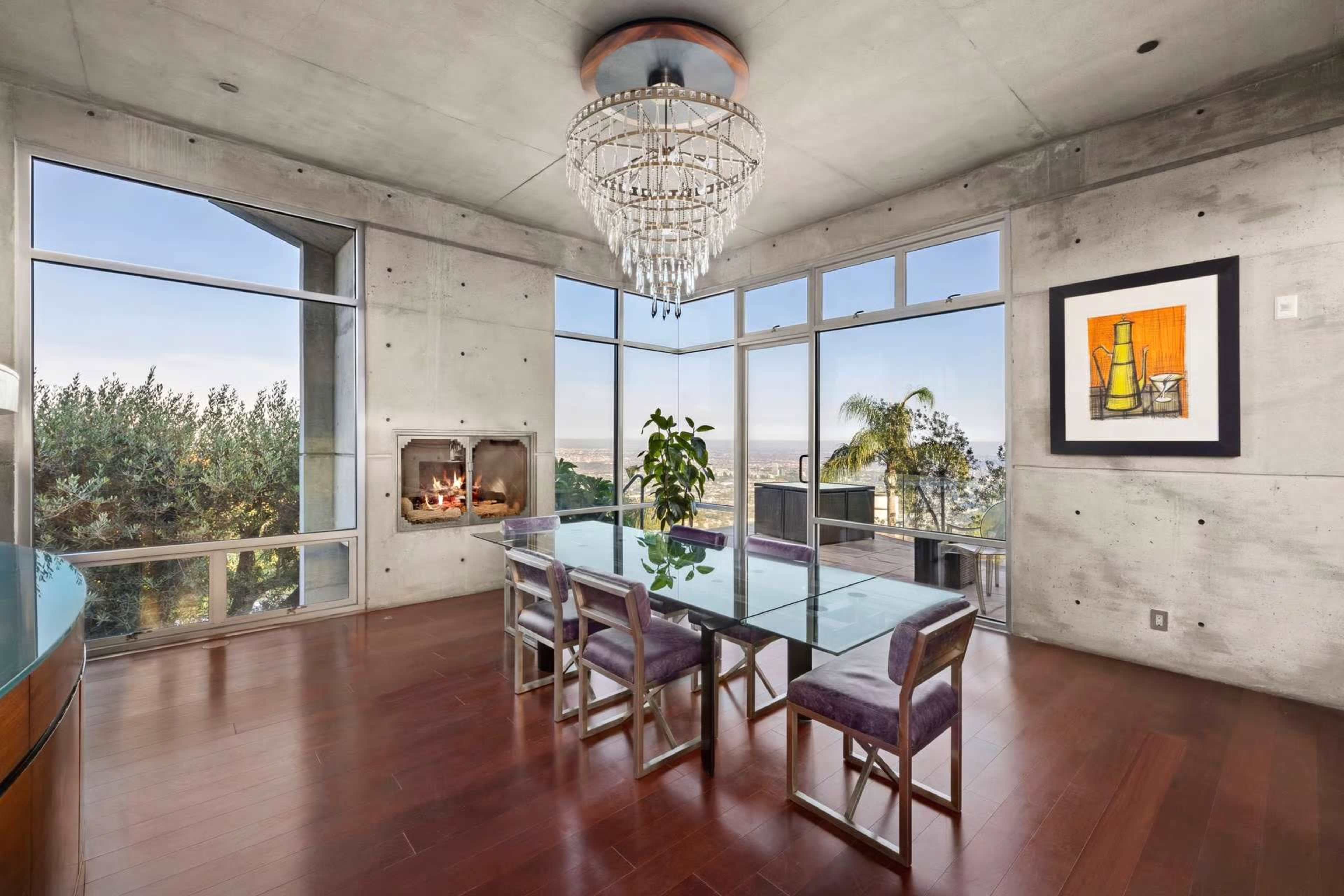 The image shows a modern dining area with a glass table, upholstered chairs, a chandelier, and large windows that offer a view of the outdoors.