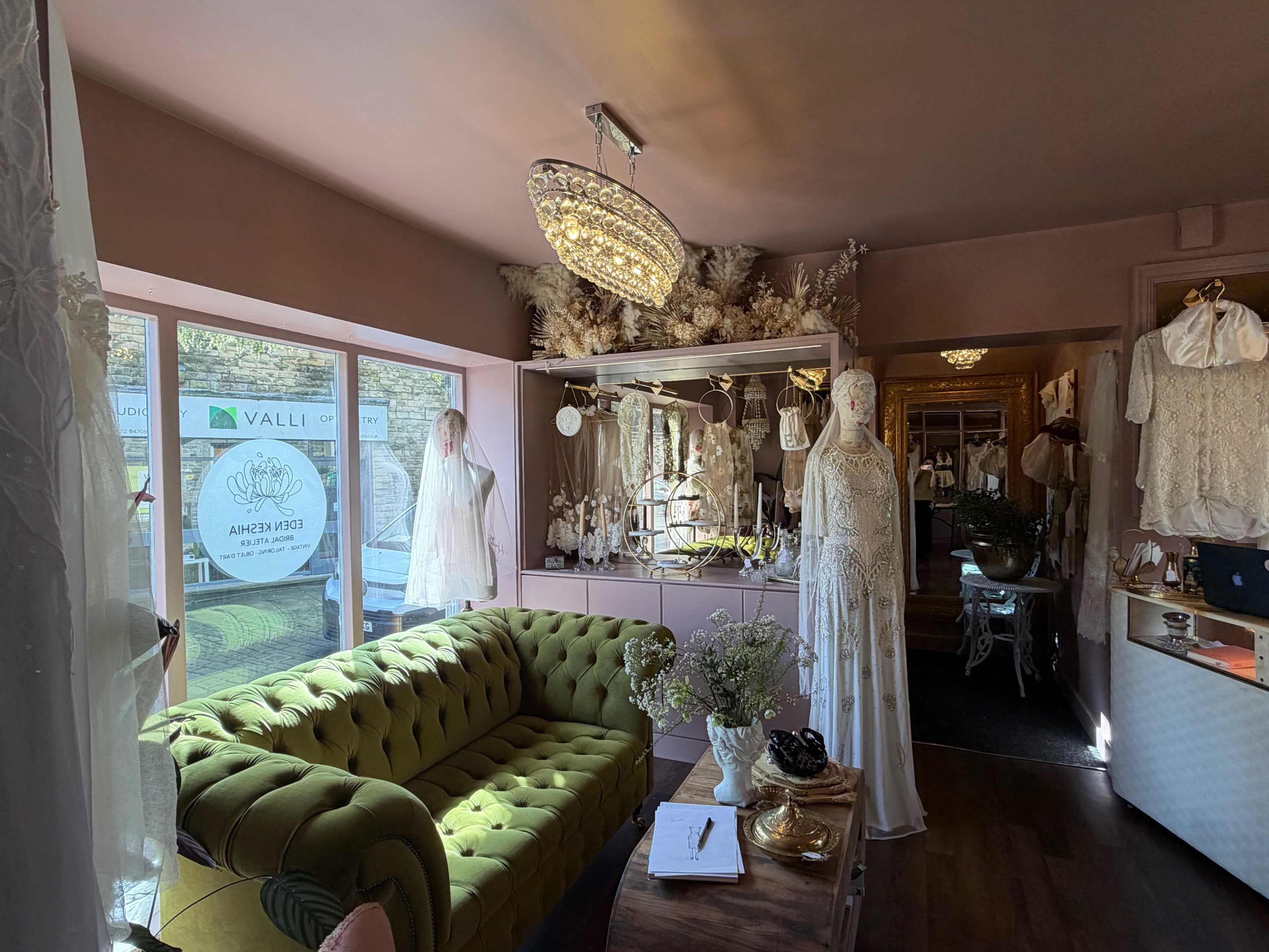 The image shows a stylish boutique interior featuring a green velvet sofa, display mannequins dressed in elegant garments, and a chandelier overhead.
