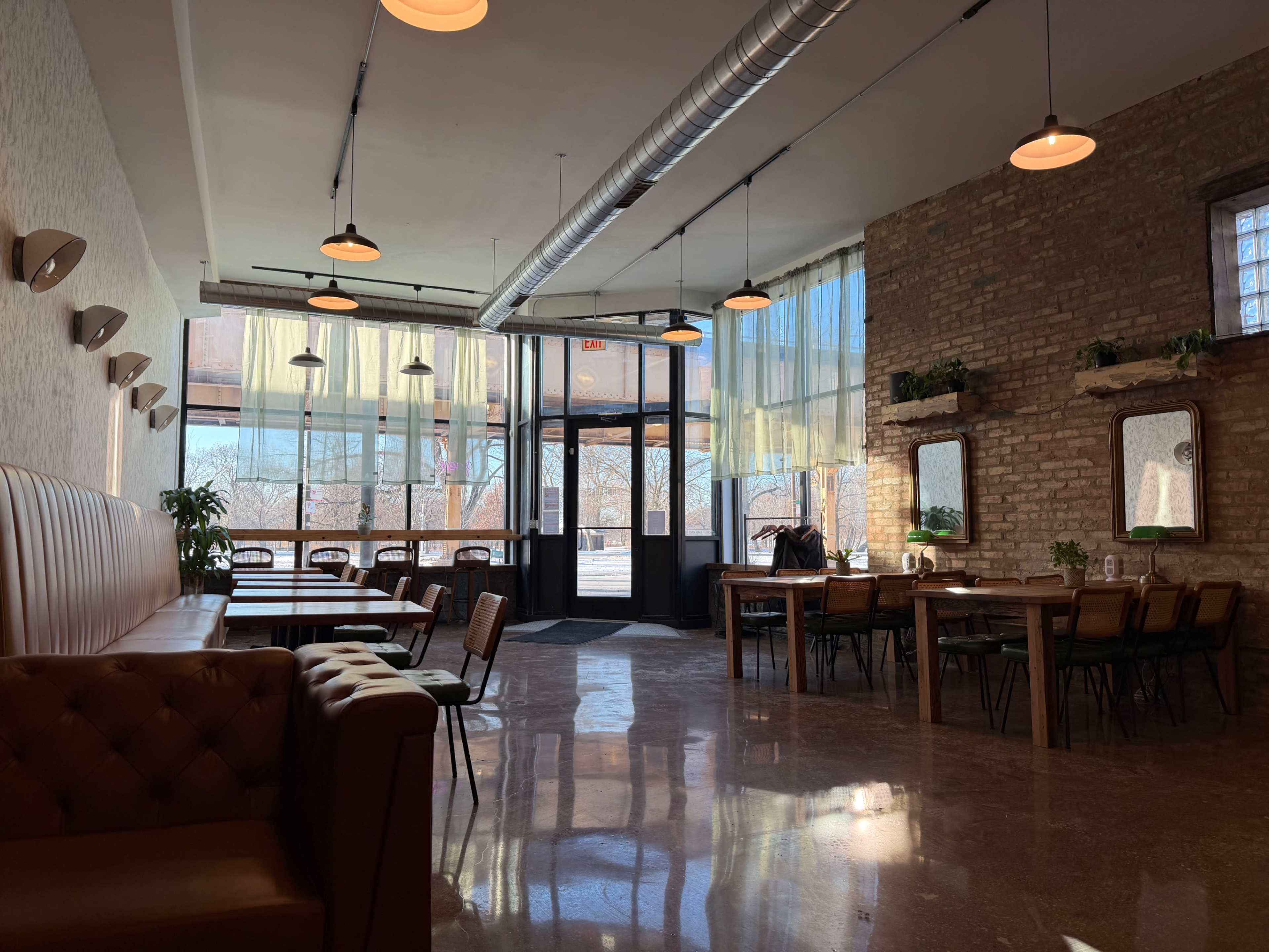 The image shows a spacious café interior featuring a mix of seating options, large windows allowing natural light in, and exposed brick walls.