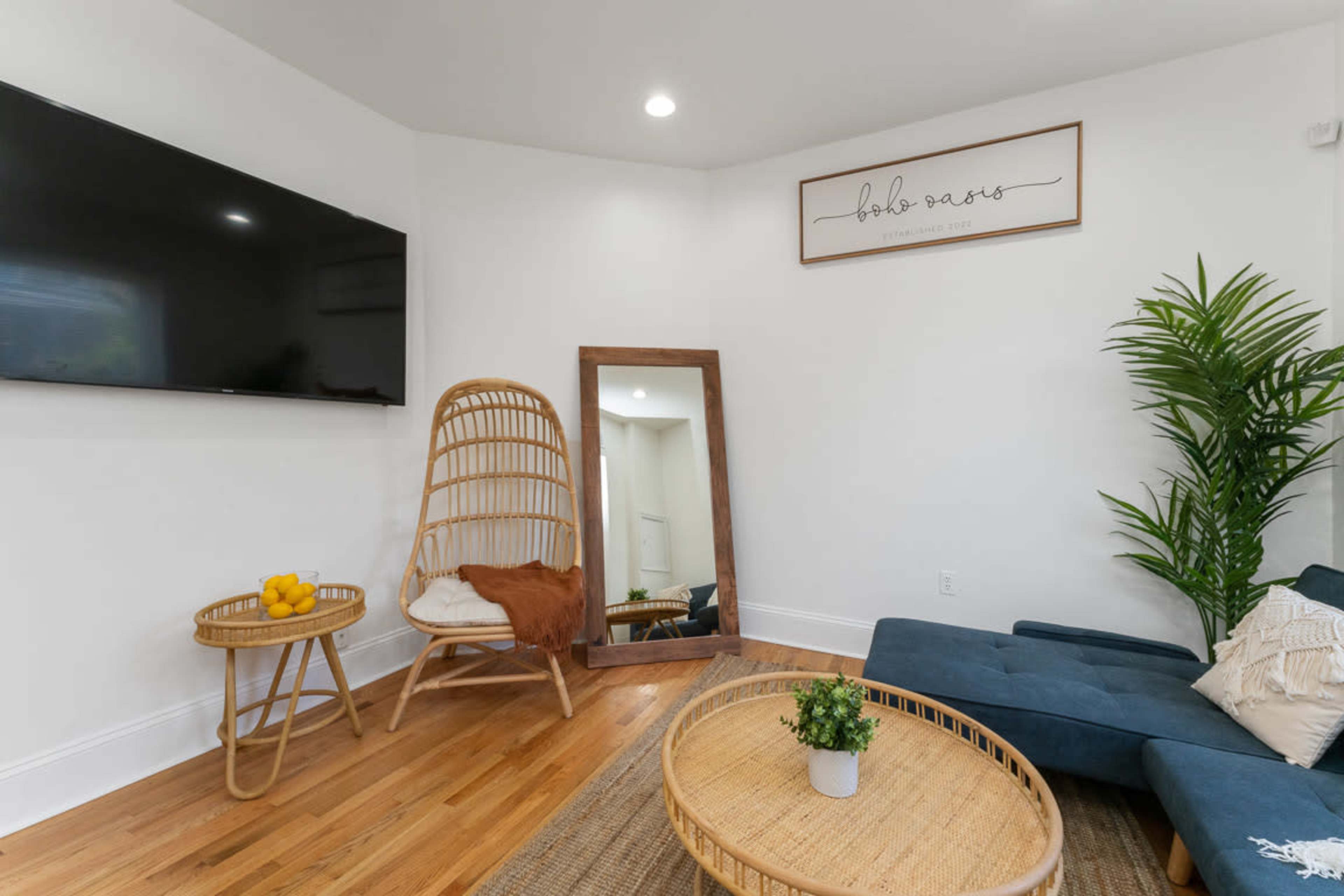 A modern living room with a large wall-mounted television, a rattan chair, a mirror, a coffee table, and green plants.