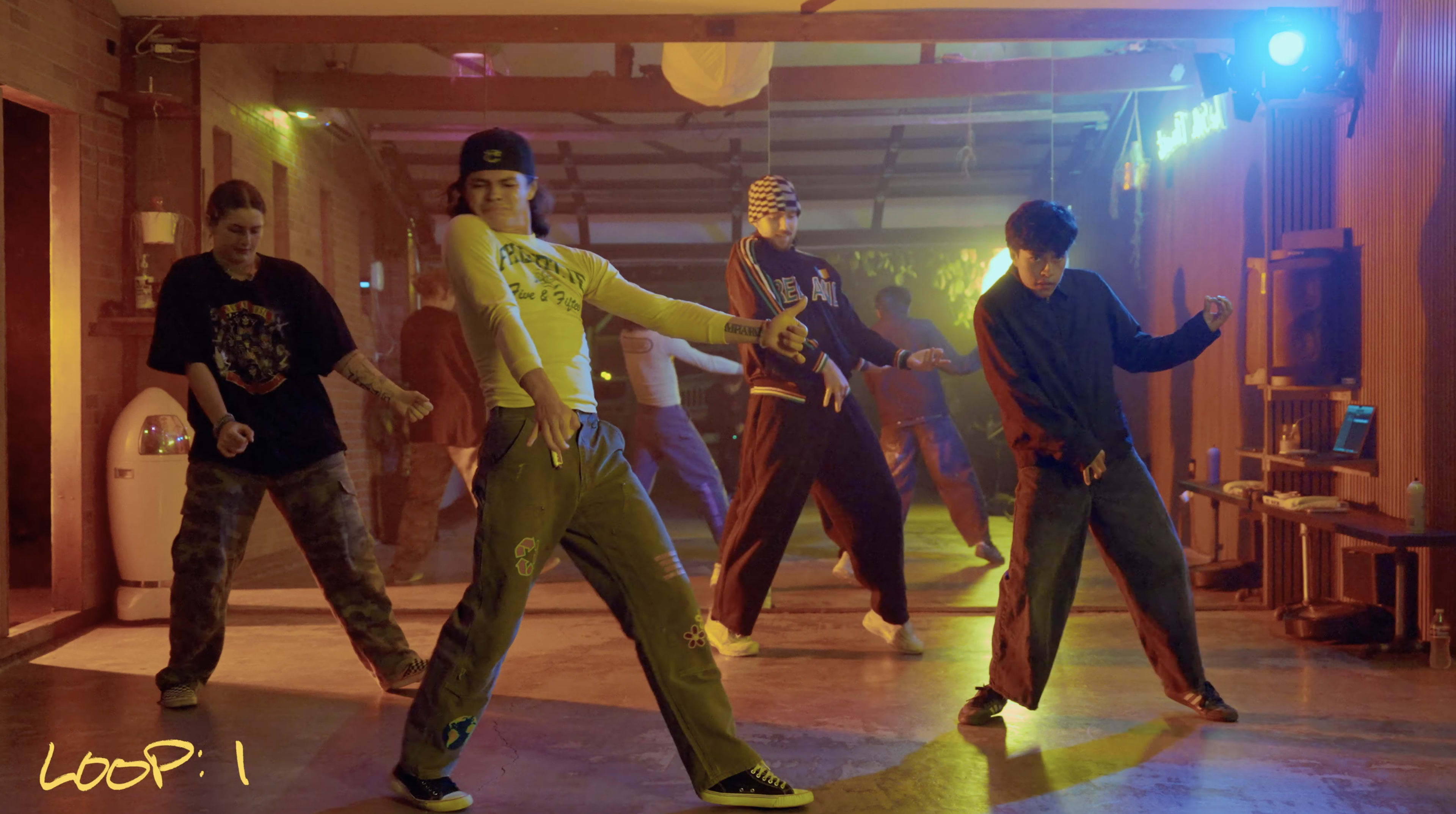 A group of dancers perform in a lively studio, illuminated by colorful lights, showcasing a variety of styles and movements.