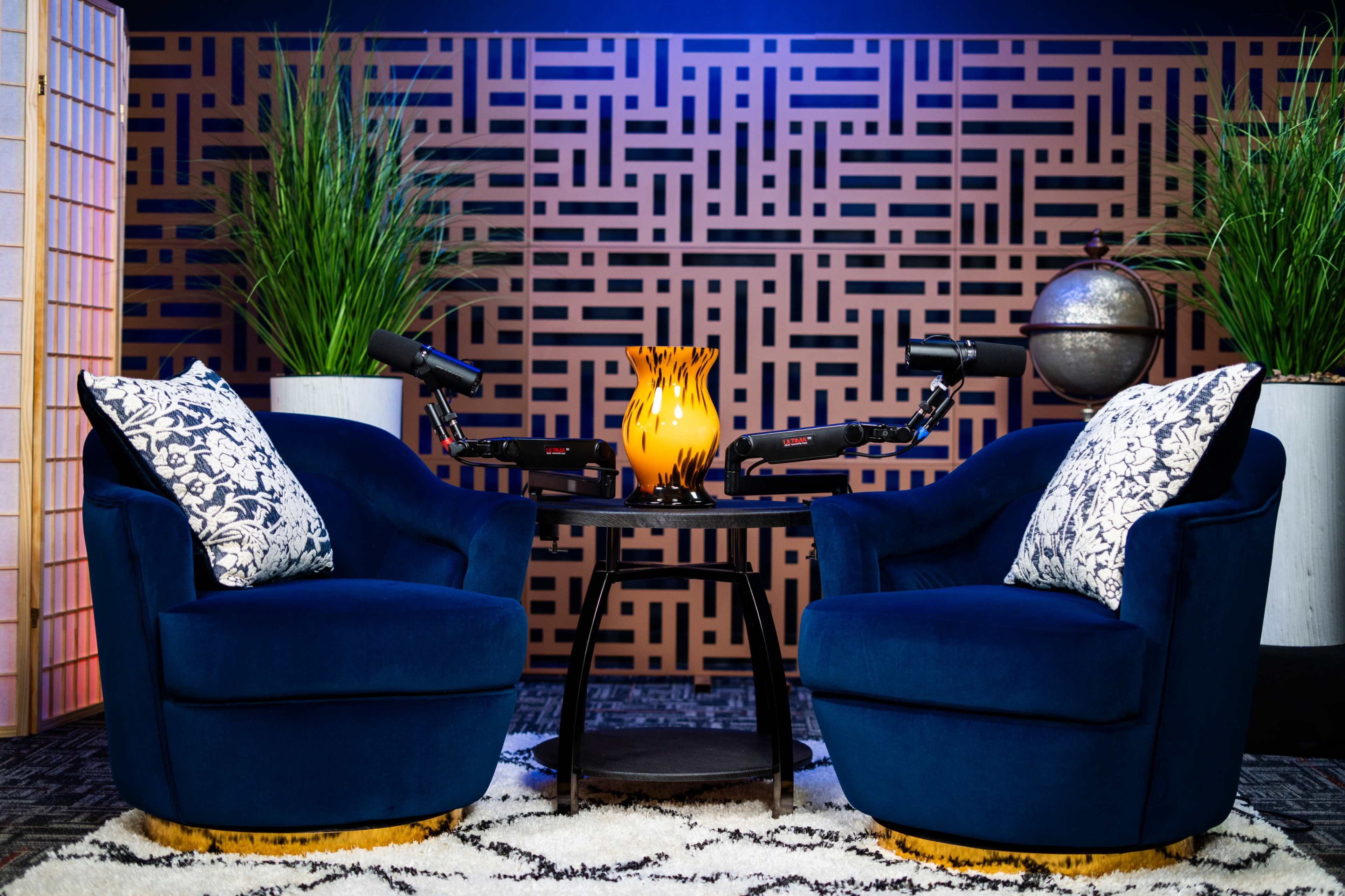 The image features two blue swivel chairs with decorative pillows, positioned around a small black table with a lit decorative vase, all set against a patterned wall and surrounded by potted plants.