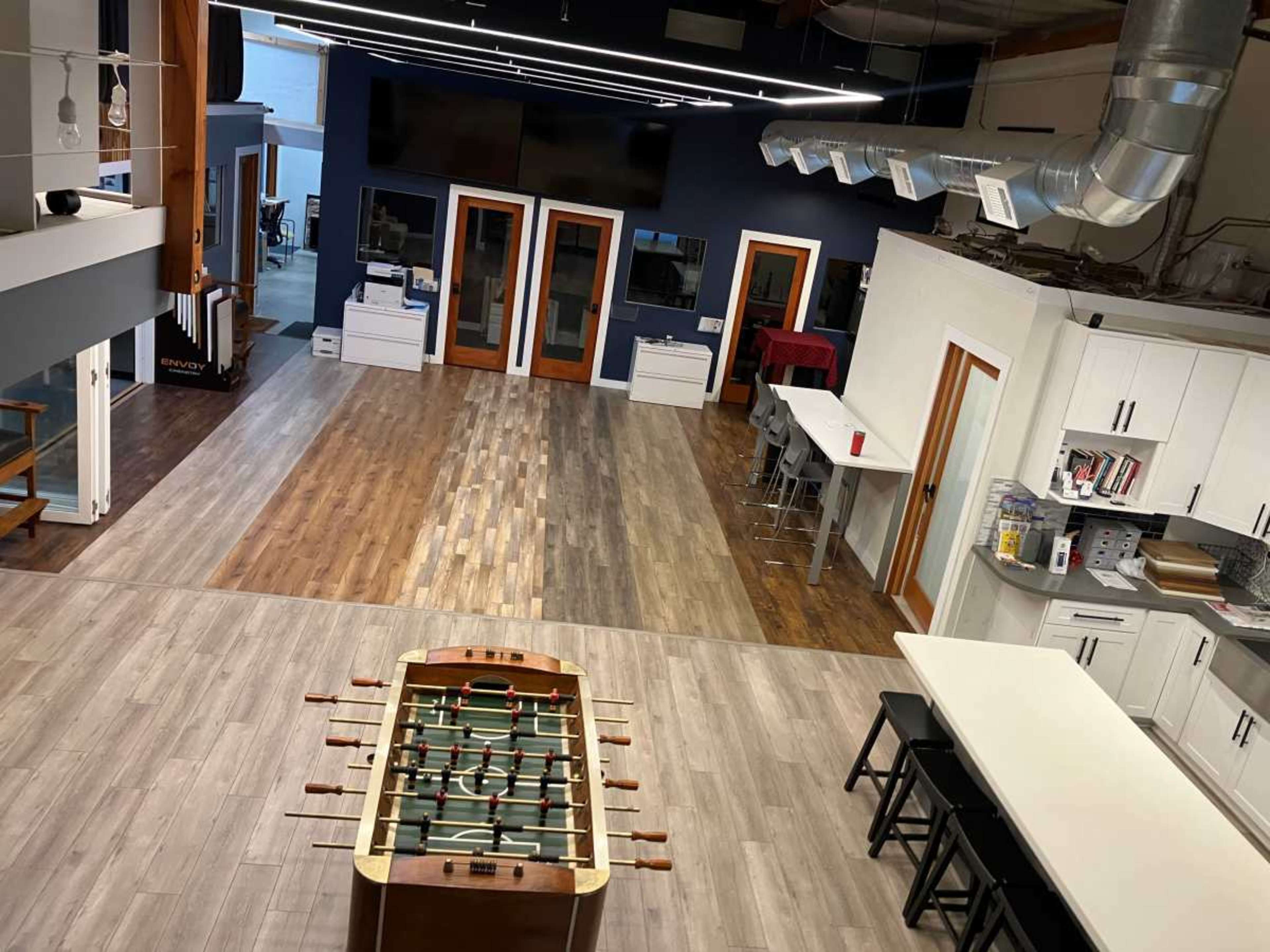 The image shows a spacious indoor recreation area featuring a foosball table, various seating arrangements, and multiple workstations.