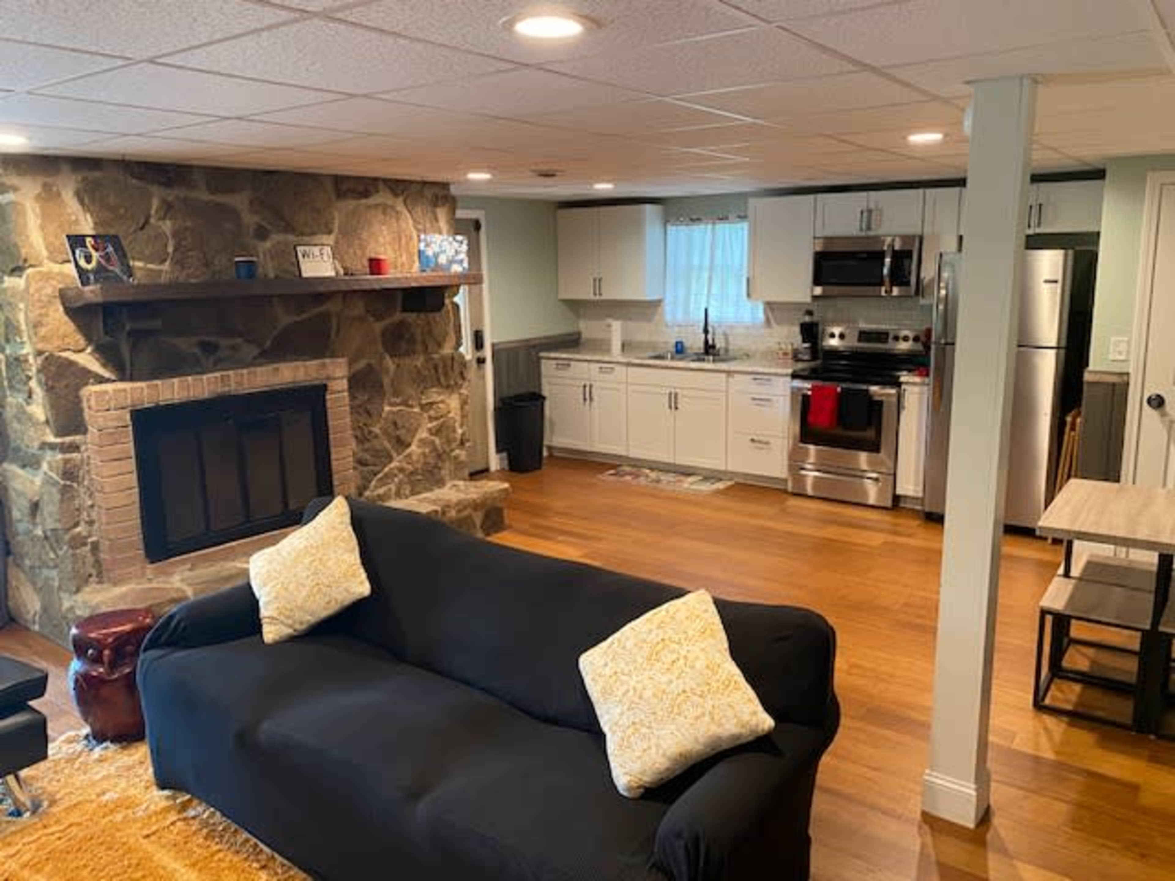 A cozy living space with a black couch, a stone fireplace, and an adjacent kitchen featuring stainless steel appliances and white cabinets.