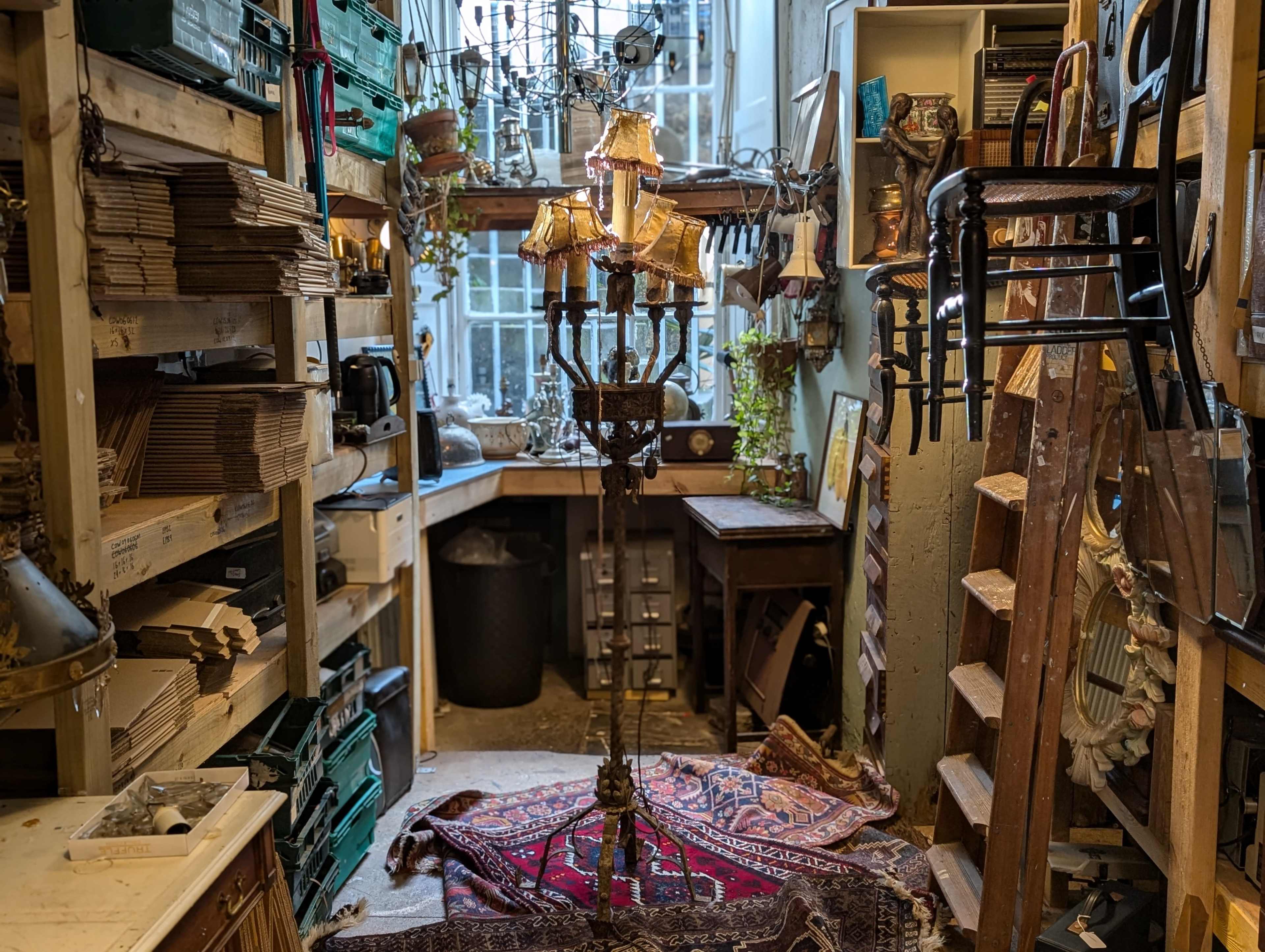The image shows a small, cluttered workshop filled with shelves of materials, tools, and a central standing lamp surrounded by a patterned rug.