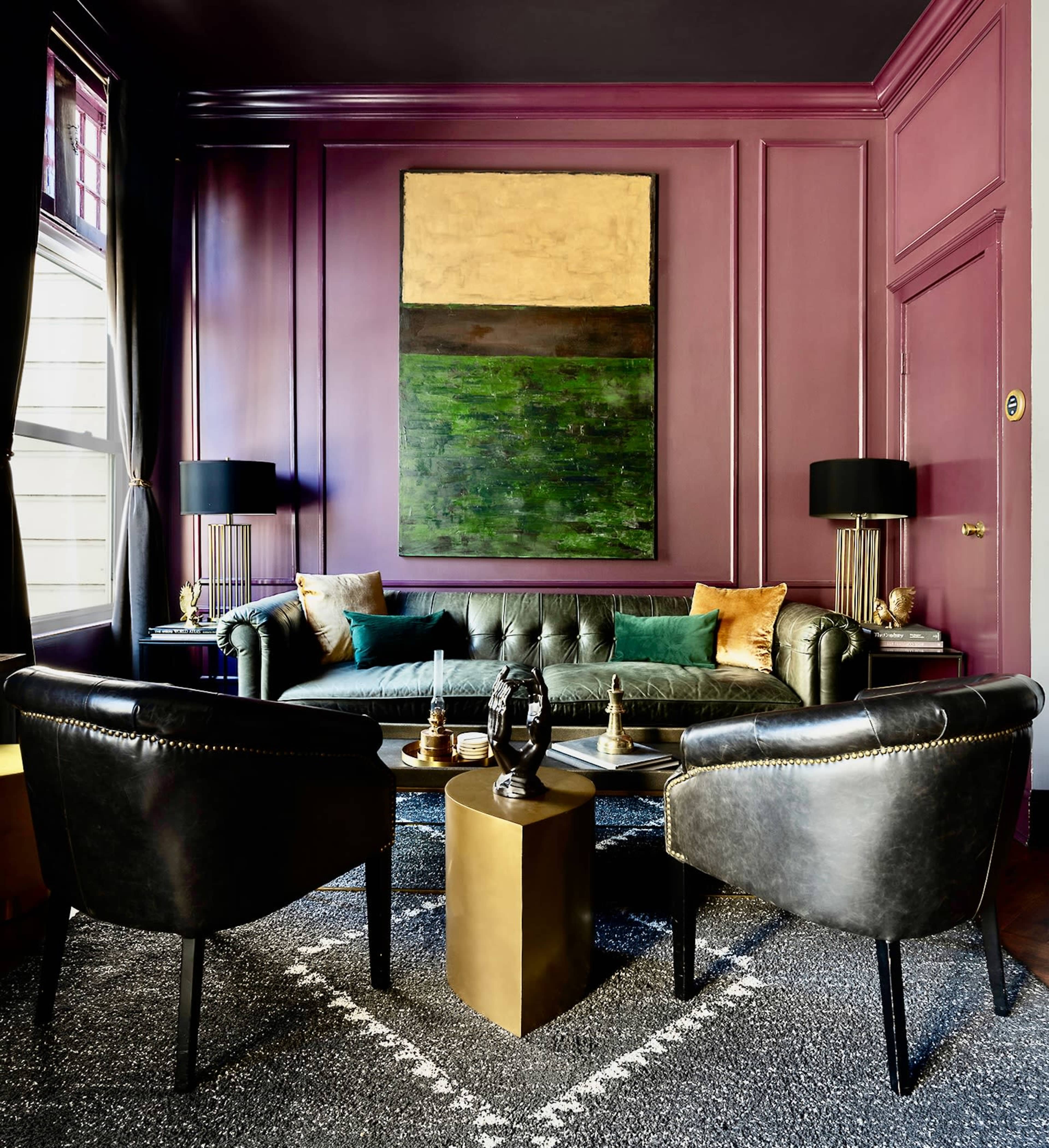 A stylish sitting area with a green leather couch, two black chairs, and a large abstract painting on a purple wall.