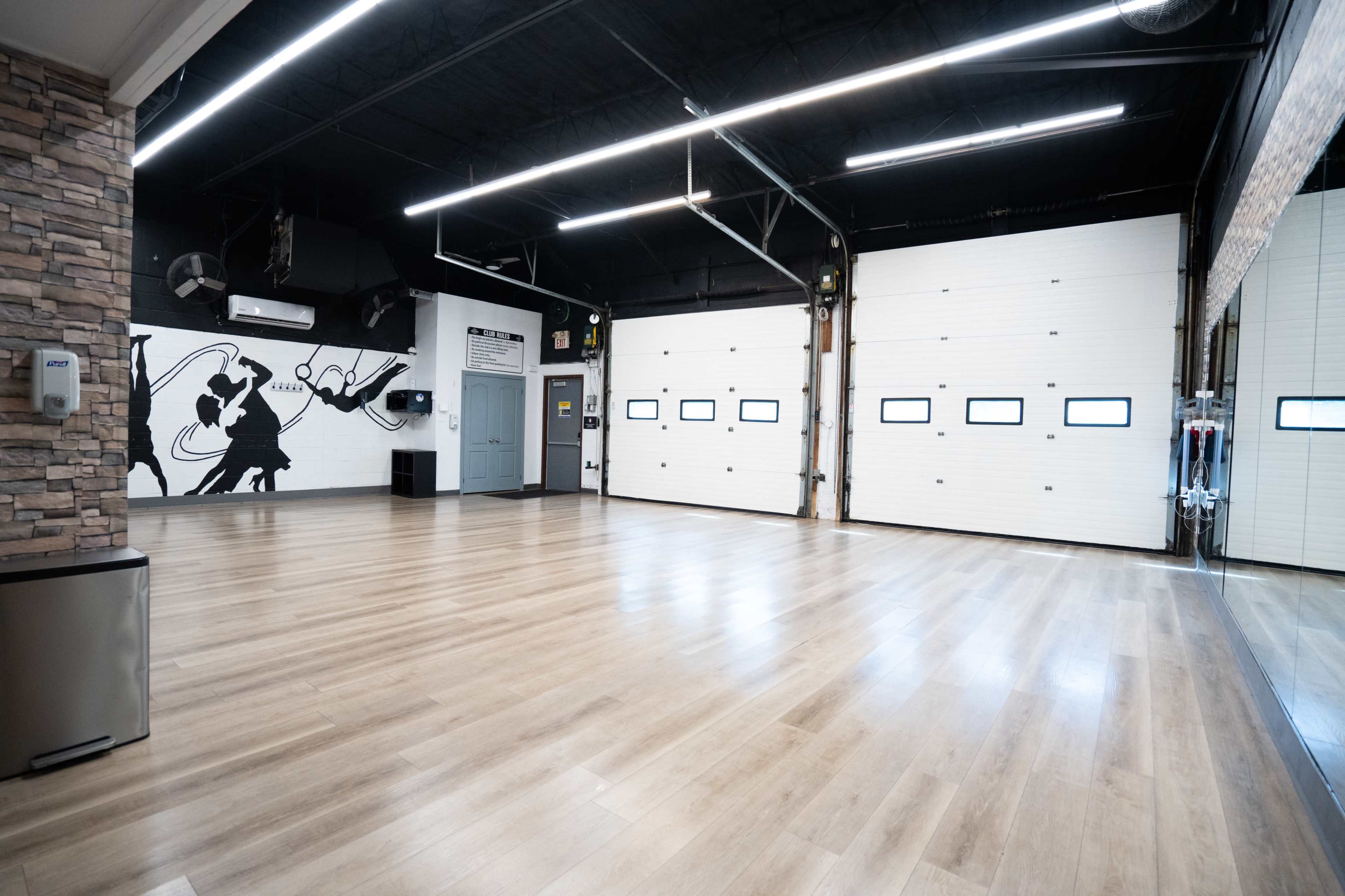 The image shows a spacious room with a hardwood floor, large garage doors, a mirrored wall, and a mural depicting a musical scene.