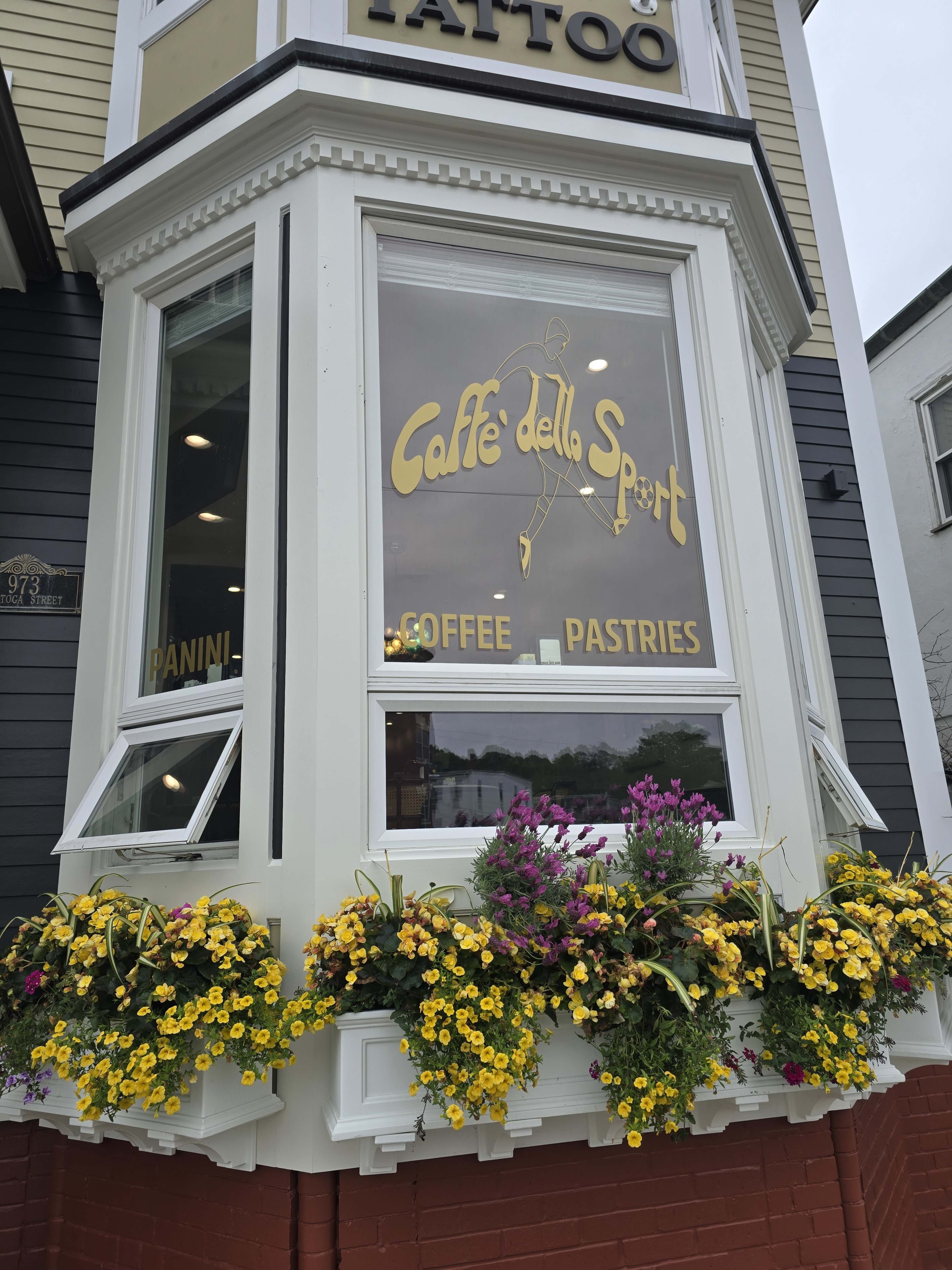 The image shows a café storefront with large windows displaying the name "Caffè della Sport" along with signs for panini, coffee, and pastries, accented by colorful flower boxes underneath.