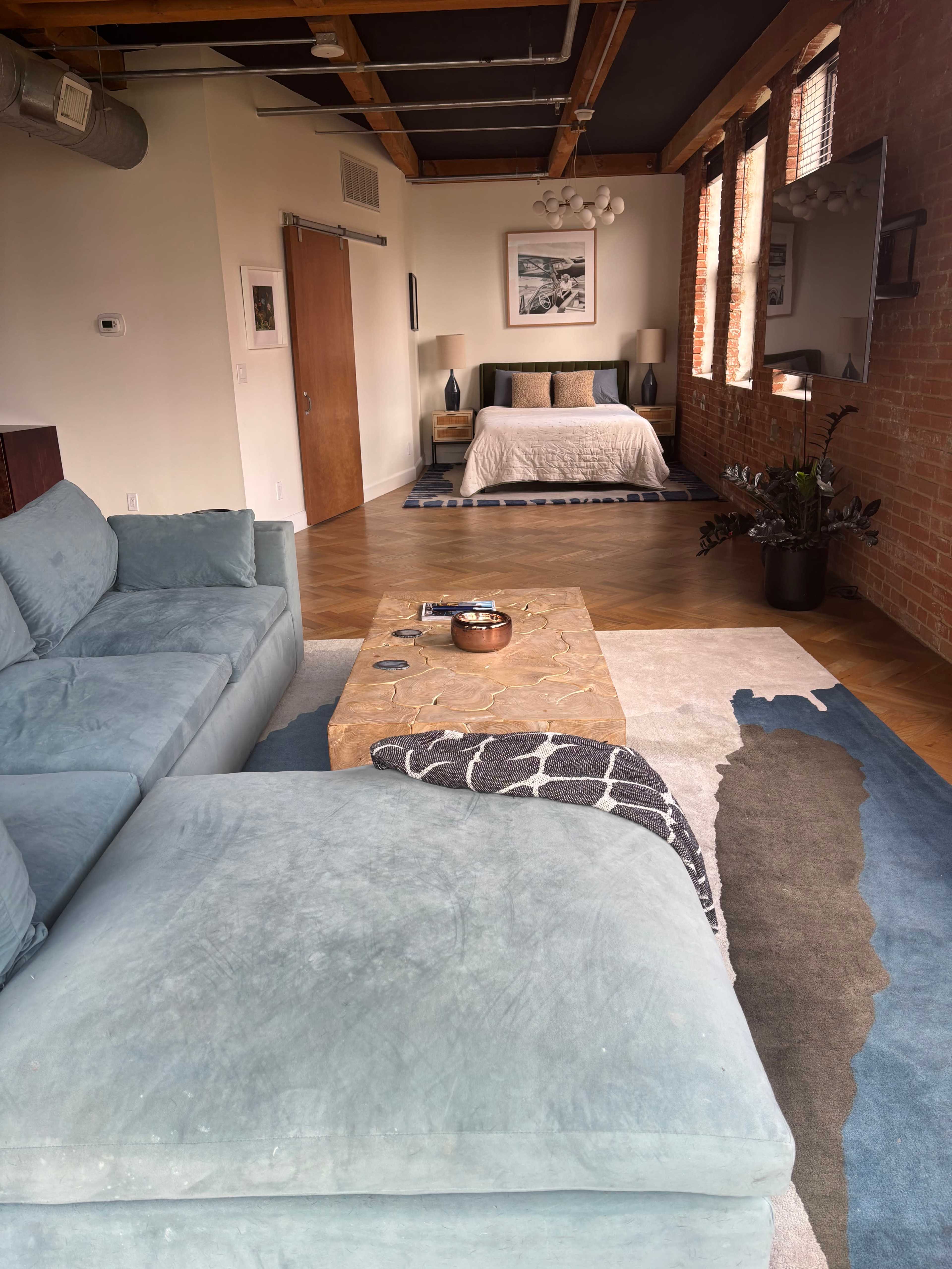The image shows a spacious modern bedroom and living area with a large sofa, a coffee table, and a bed against a wall of exposed brick.