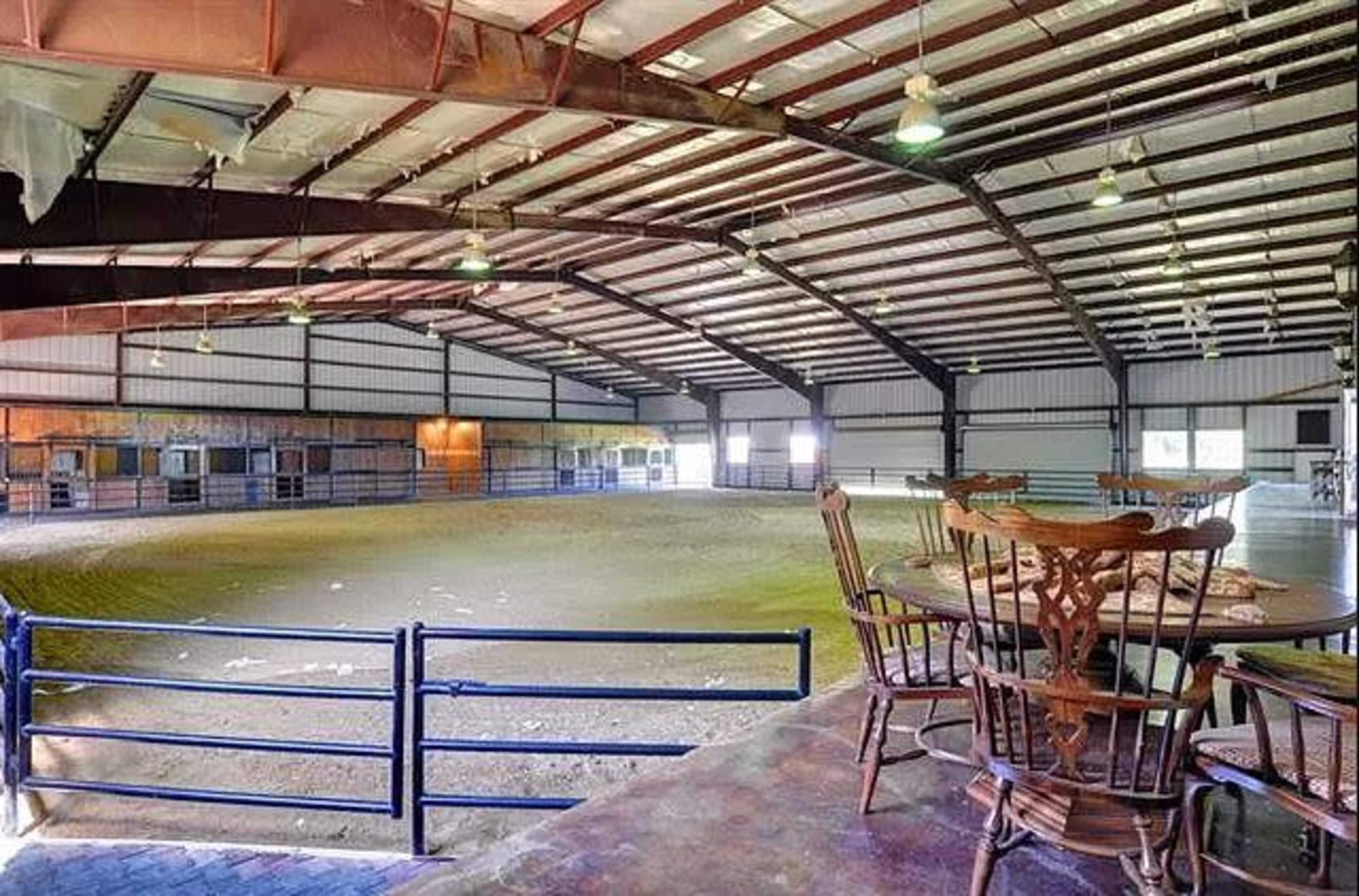 A spacious indoor riding arena with a dirt floor and wooden furniture arranged near a railing.