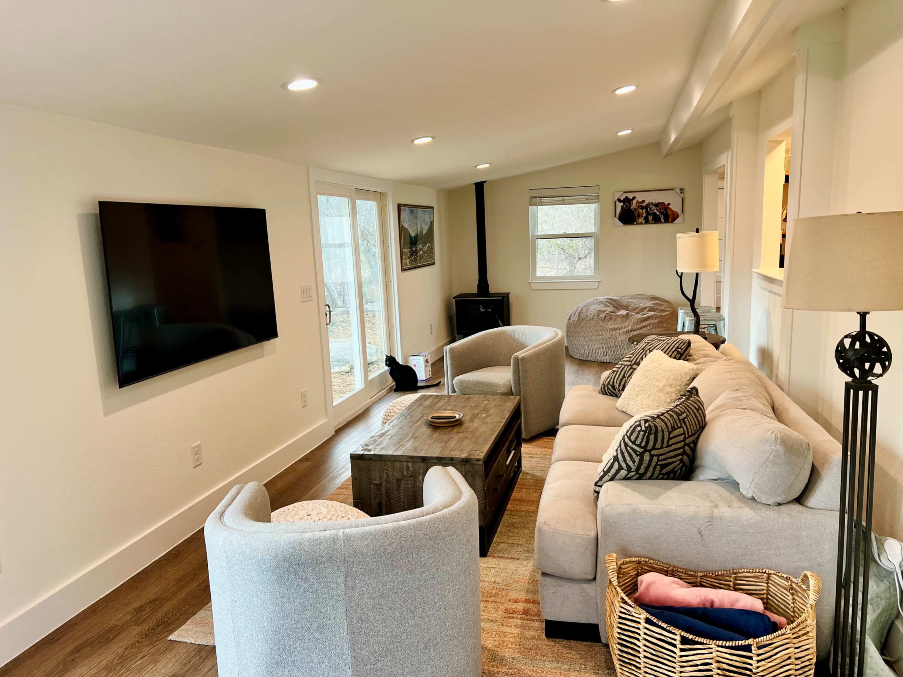 The image shows a cozy living room with a sectional sofa, two armchairs, a coffee table, and a television mounted on the wall, with large windows letting in natural light.