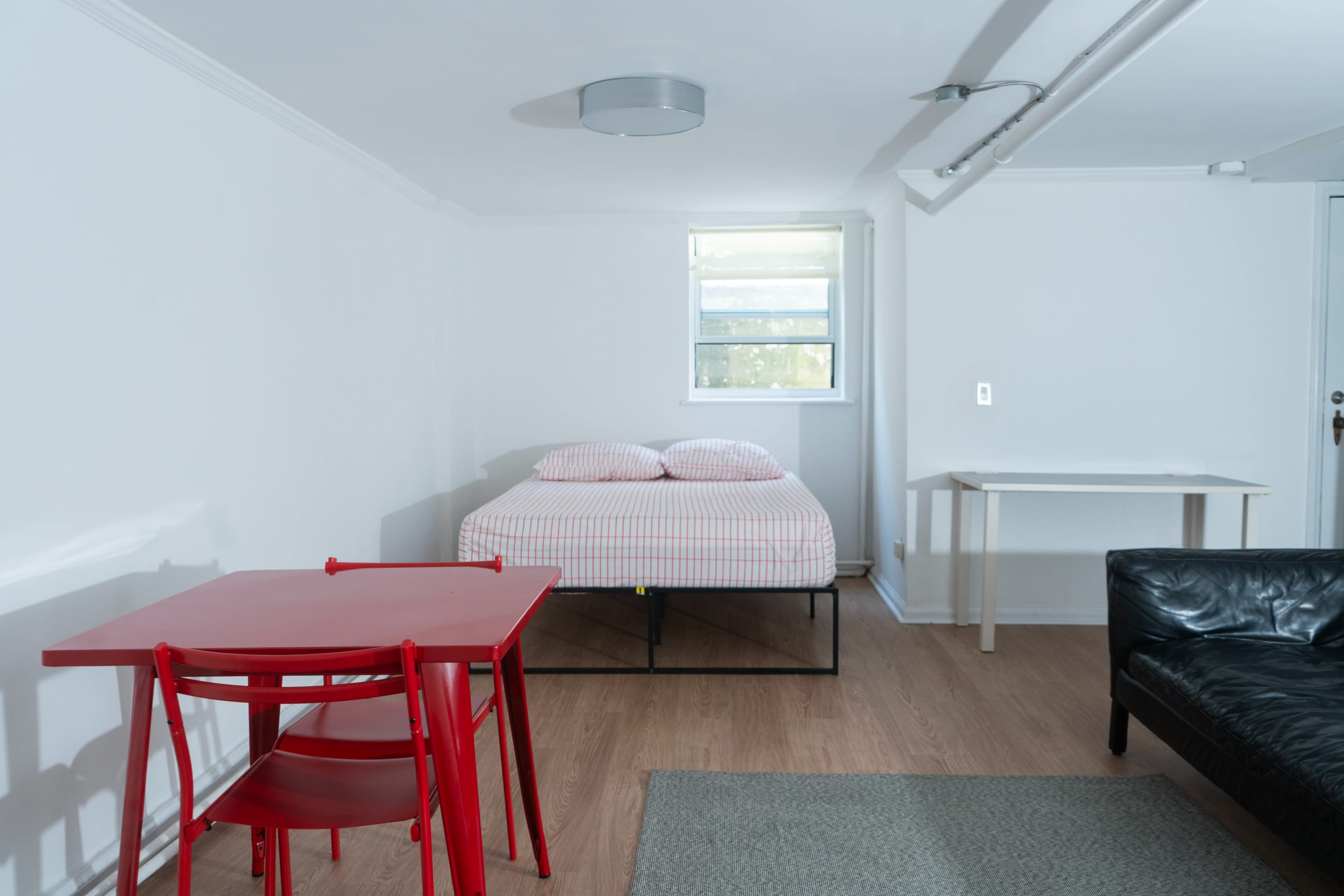 A small, minimalist room features a bed with a striped comforter, a red table with two chairs, and a black couch, all set against white walls and wooden flooring.