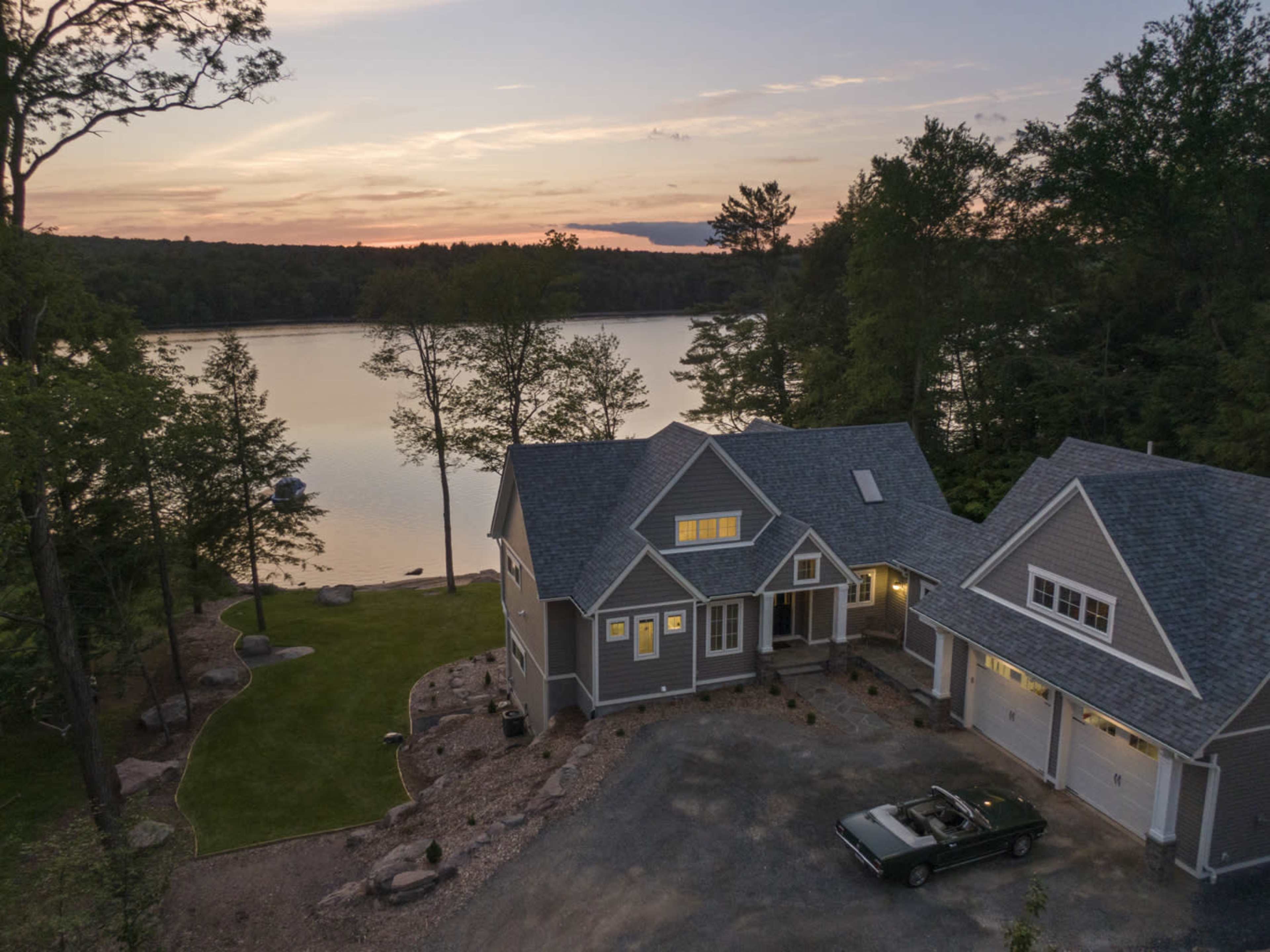 A modern house with a gravel driveway is situated near a lake, surrounded by trees, as the sun sets in the background.