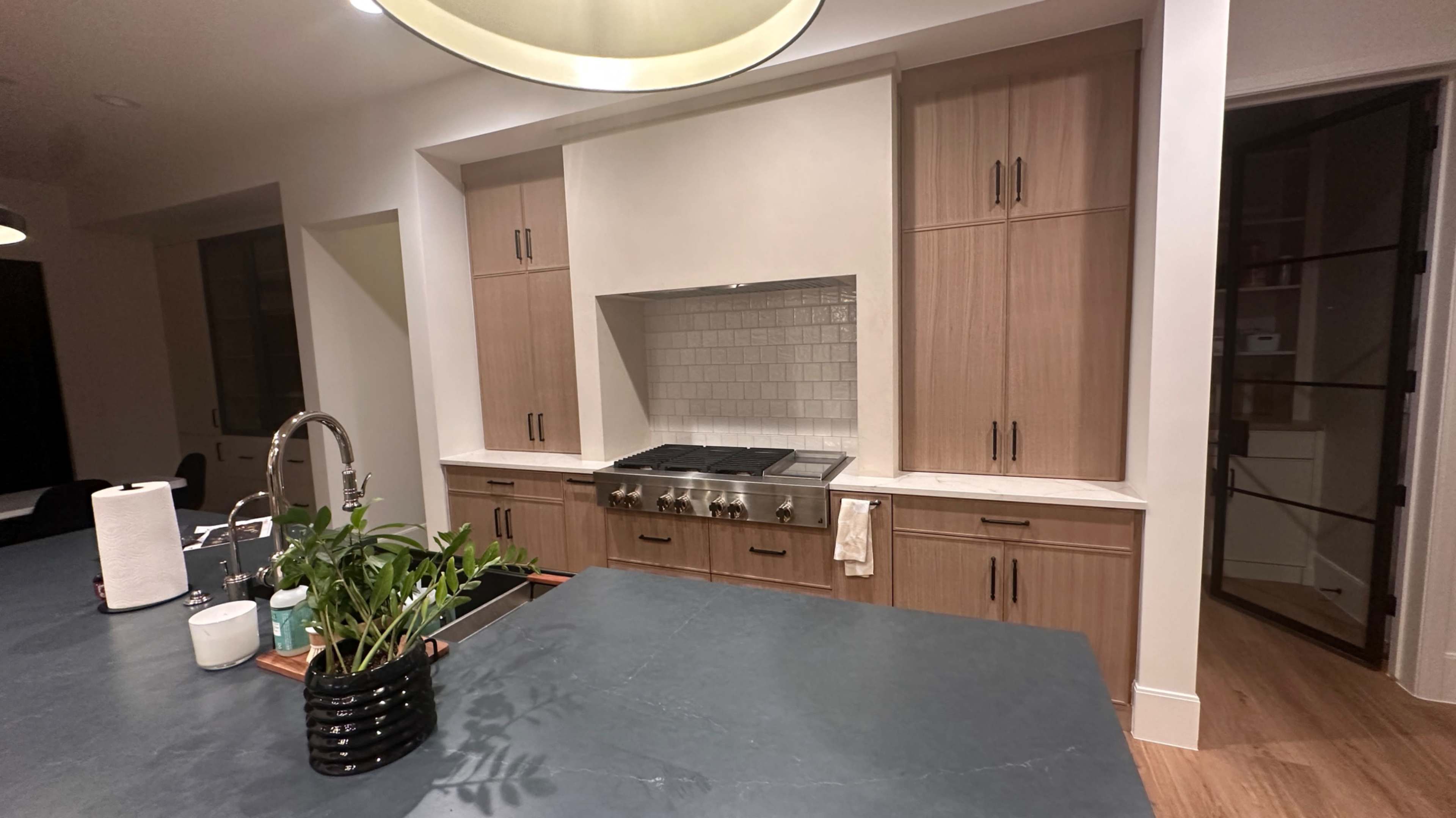 A modern kitchen featuring wooden cabinetry, a large stovetop, and a dark stone countertop with a potted plant in a black container.