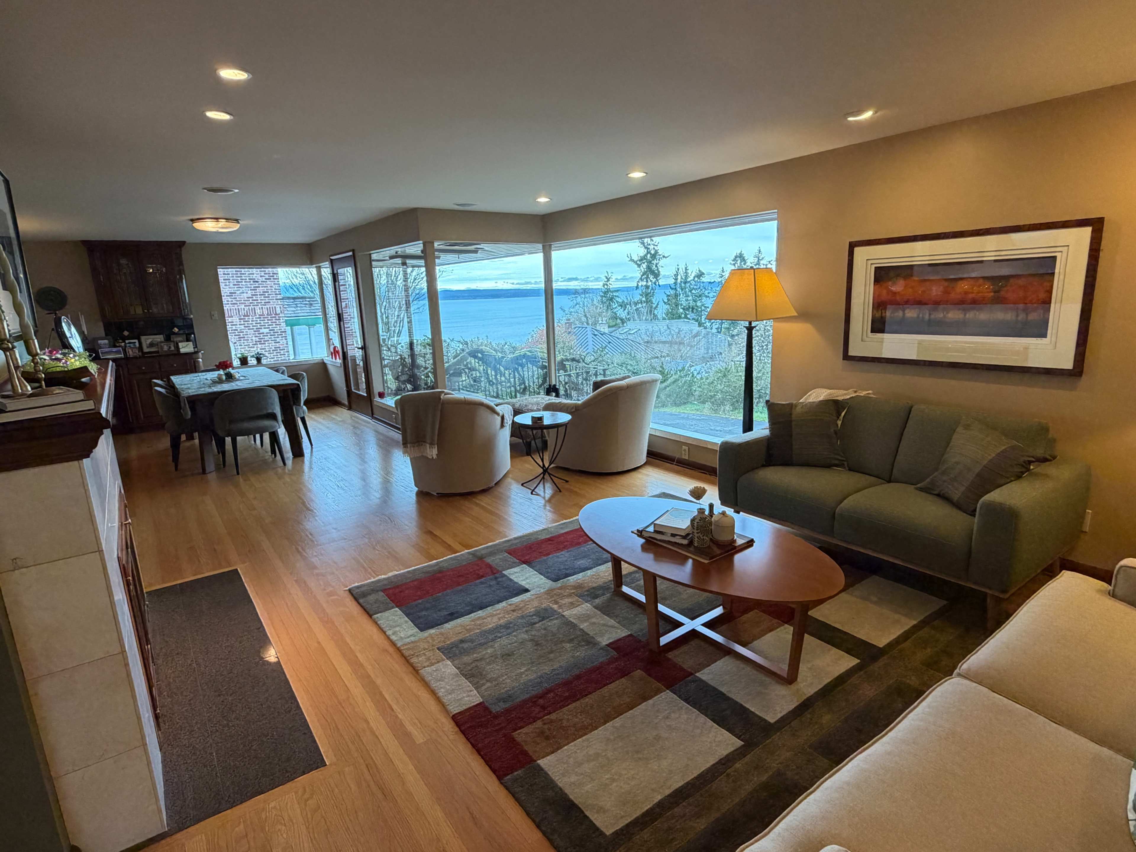The image shows a modern living room with large windows overlooking a body of water, featuring a sofa, two chairs, a coffee table, and a lamp.