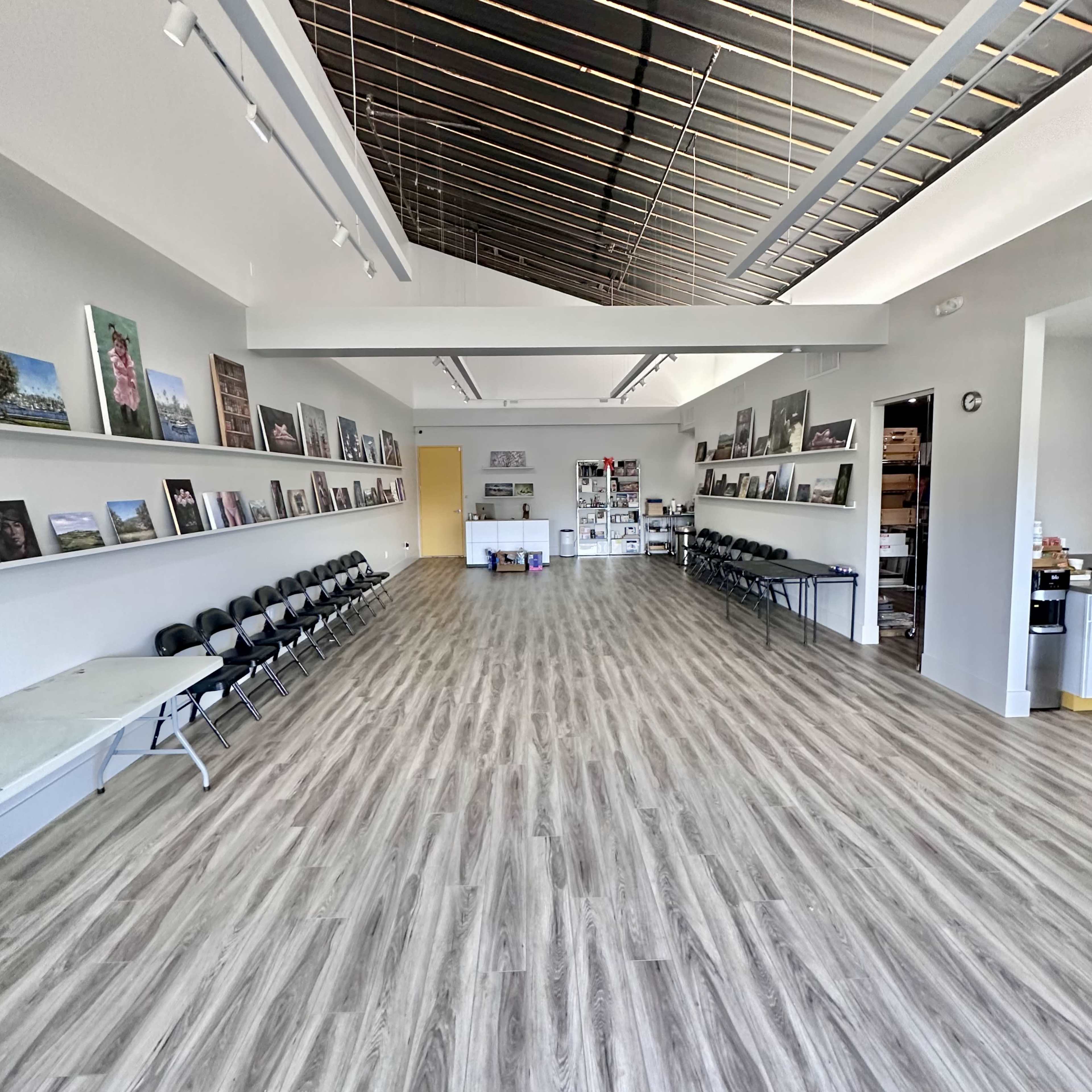 An interior space with wooden flooring, rows of folding chairs along the left wall, and framed photographs displayed on the walls.