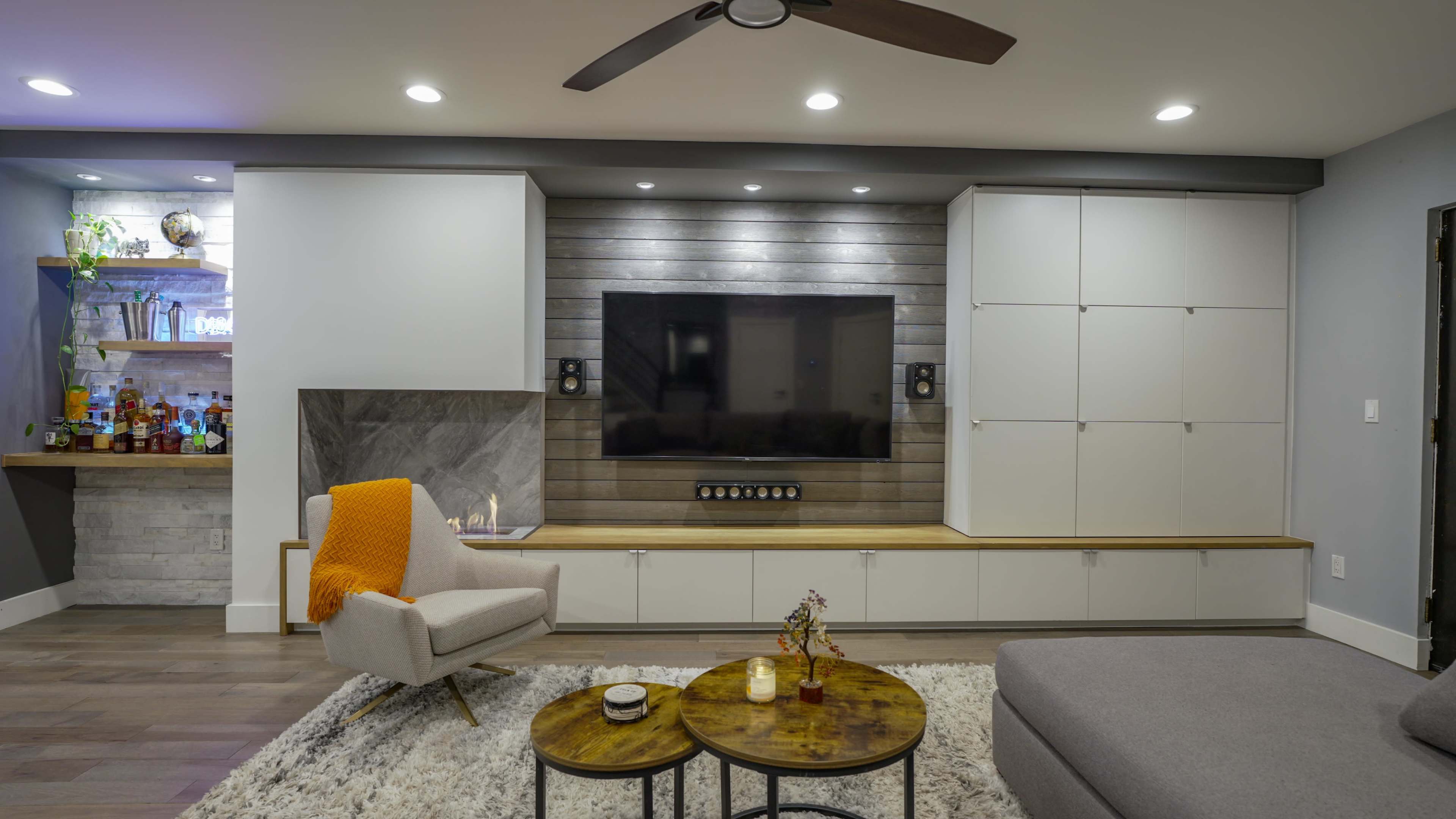The image shows a modern living room featuring a wall-mounted television, gray furniture, two circular wooden coffee tables, and a cozy armchair with an orange throw.