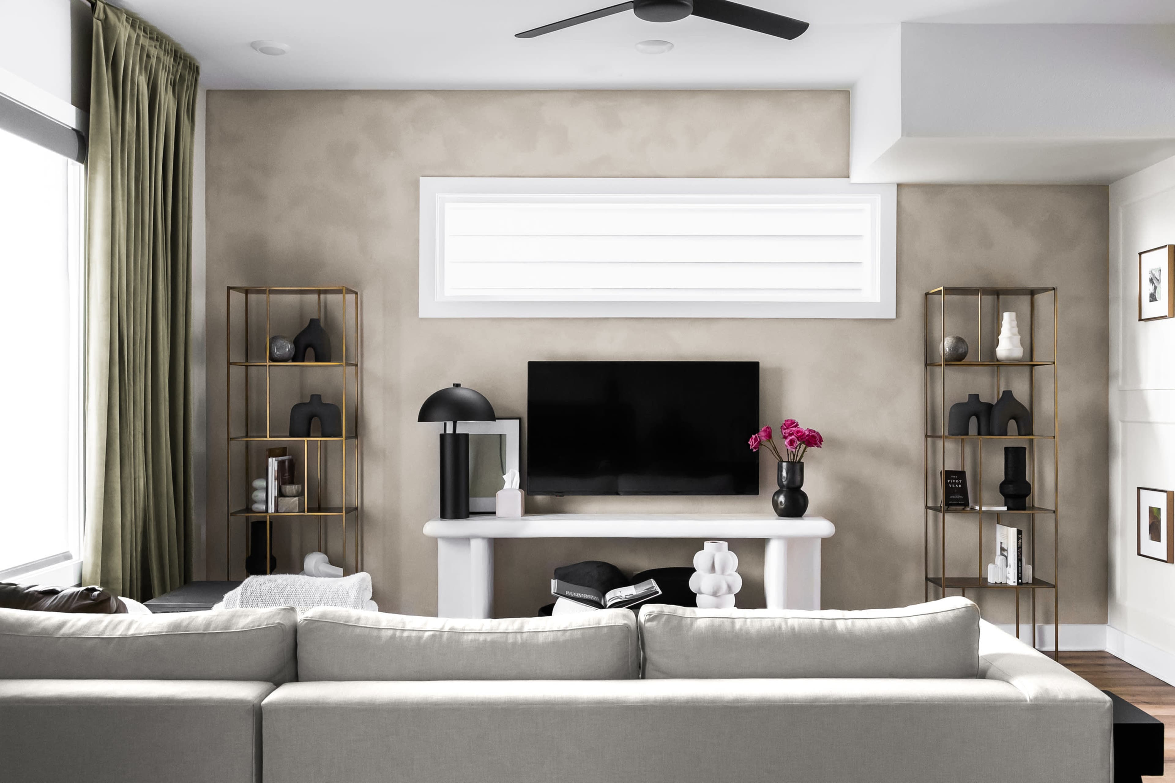 The image shows a modern living room featuring a light-colored sofa, a wall-mounted television, a decorative mantel, and gold shelving units on either side, with curtains and a few decorative items.