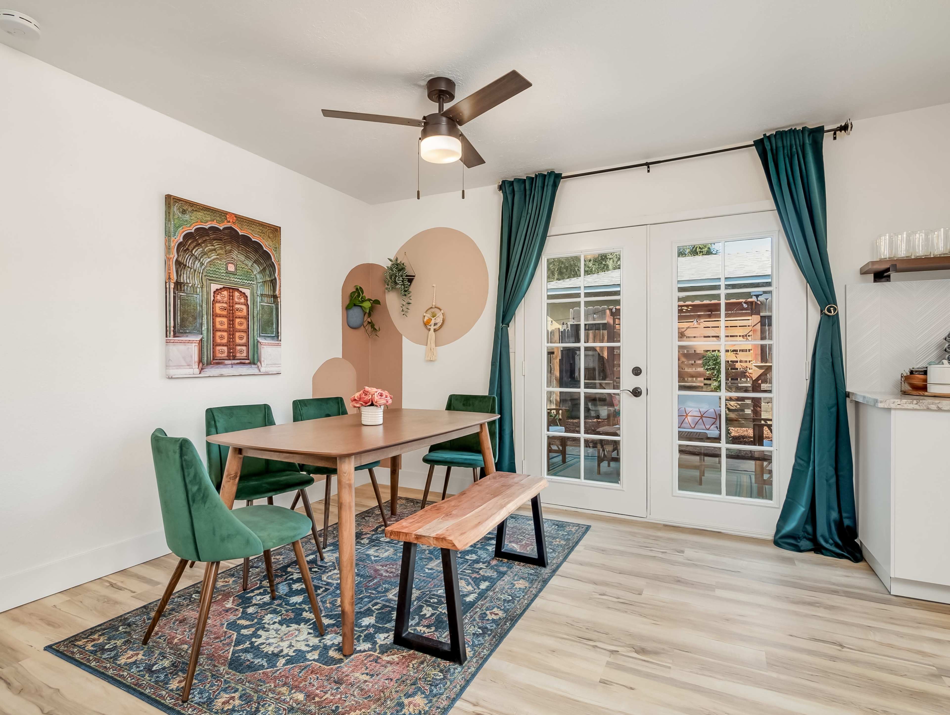 A dining area features a wooden table with green chairs, a bench, and a large artwork on the wall, illuminated by a ceiling fan and natural light from the French doors.