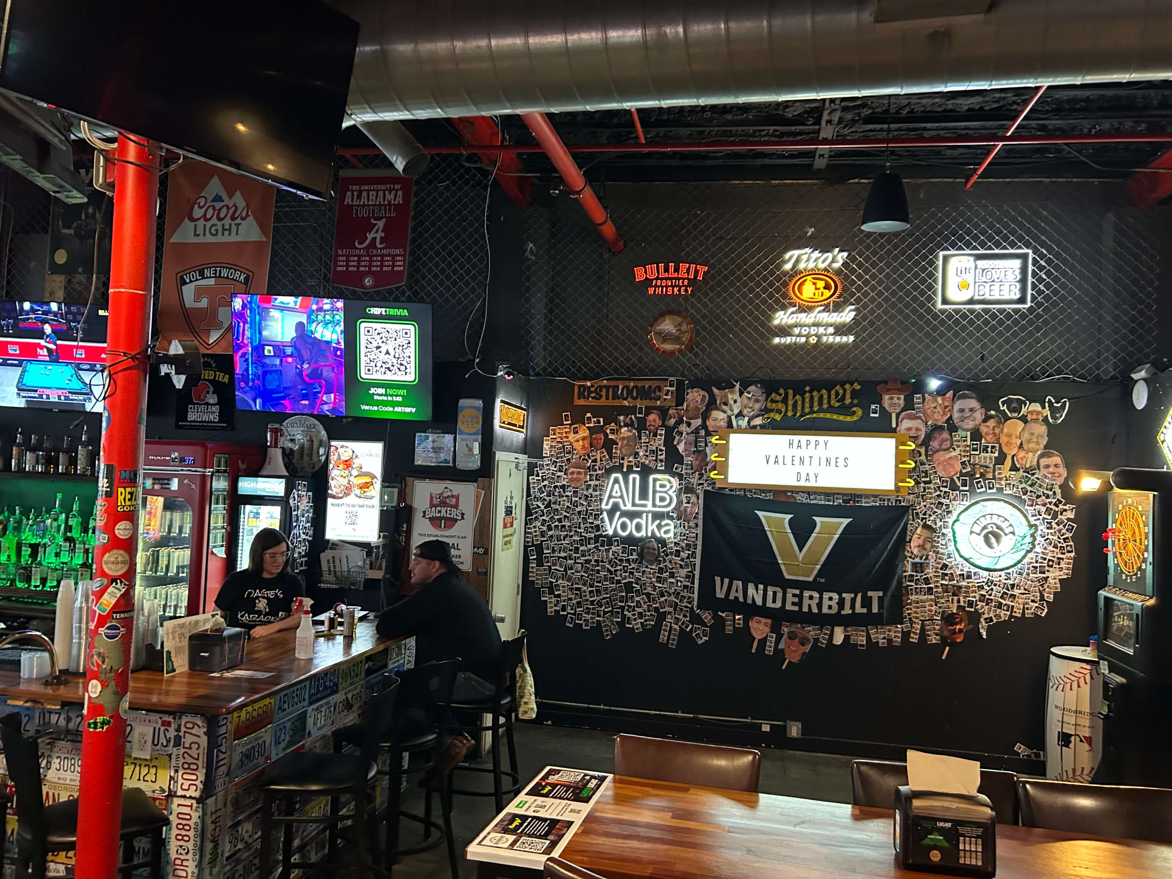 The image shows a bustling bar interior featuring a vibrant wall of decorations, illuminated signs, and patrons seated at the counter.