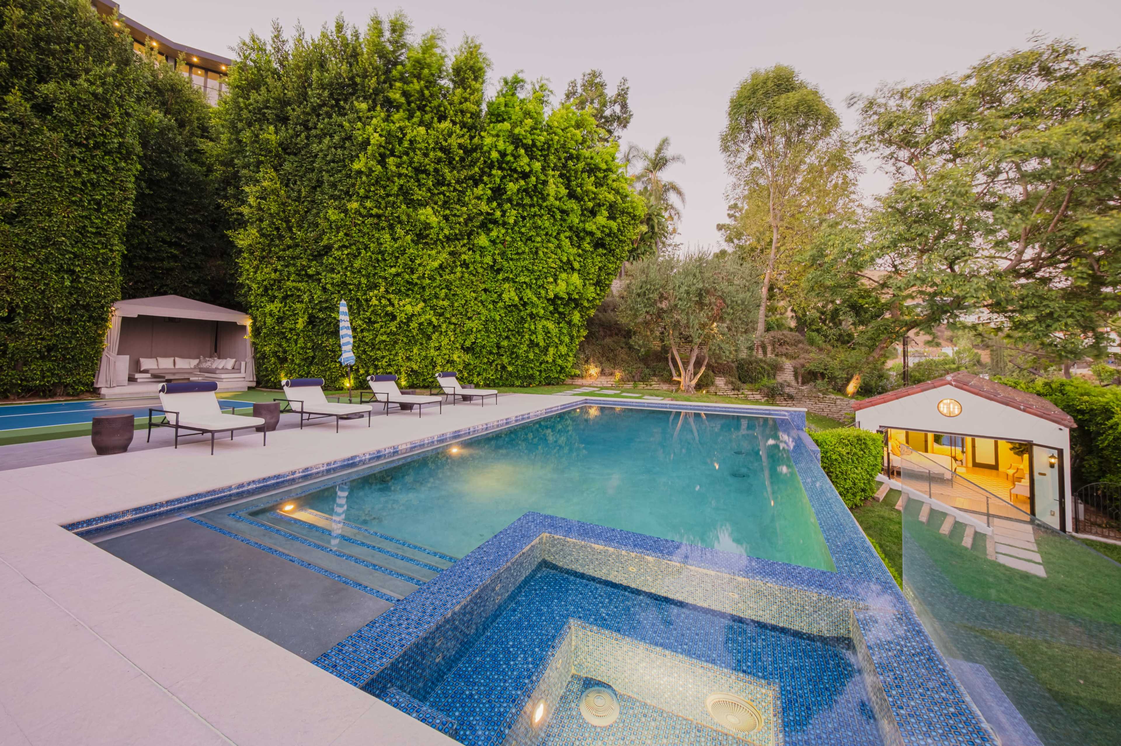 A modern backyard features a swimming pool with a spa, surrounded by lounge chairs and lush foliage.
