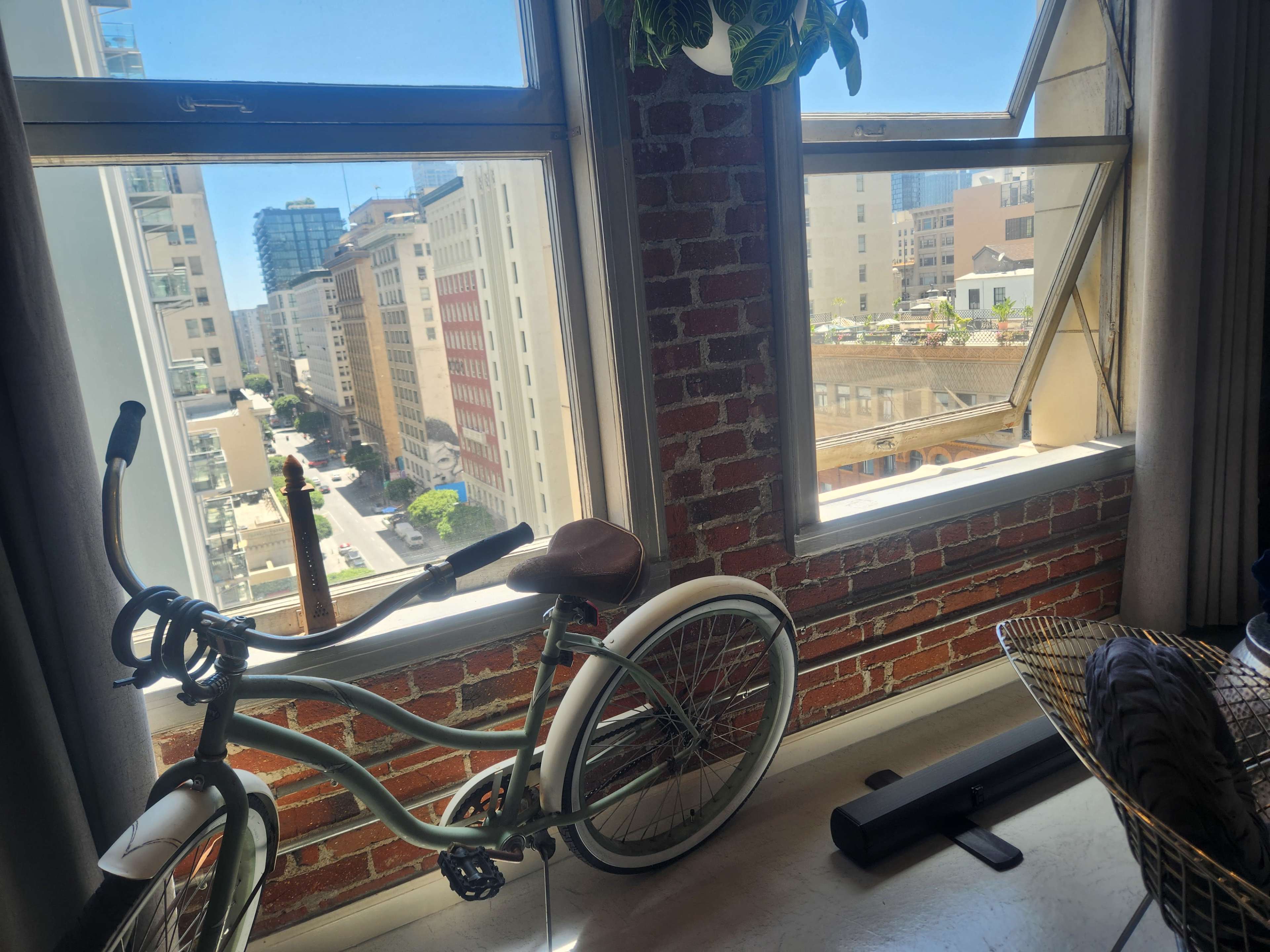 A bicycle leans against a brick wall beside a window that overlooks a city street and buildings.