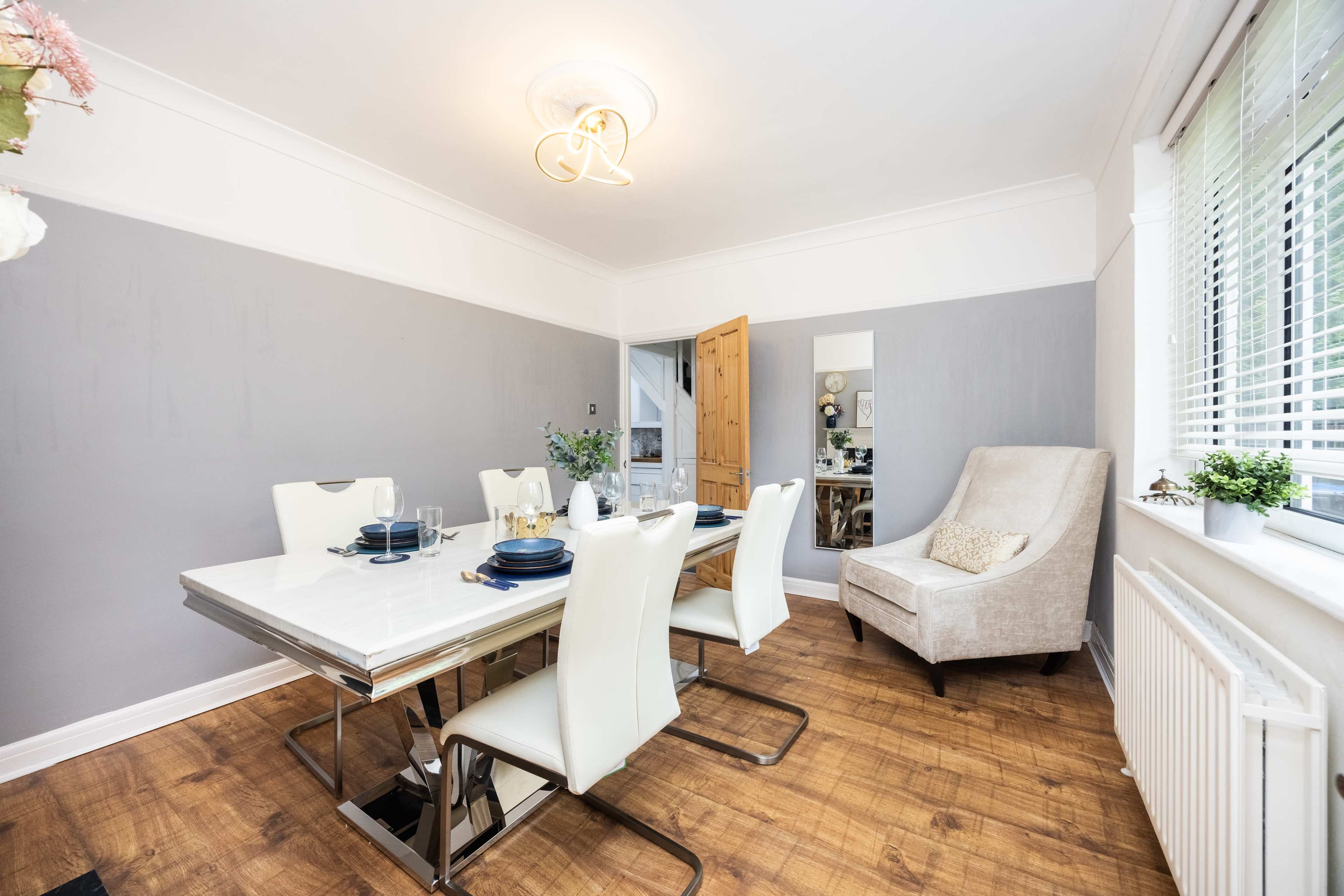 A modern dining area features a white table set for four, surrounded by sleek white chairs, with a cream armchair in the corner and a mirror reflecting the space.
