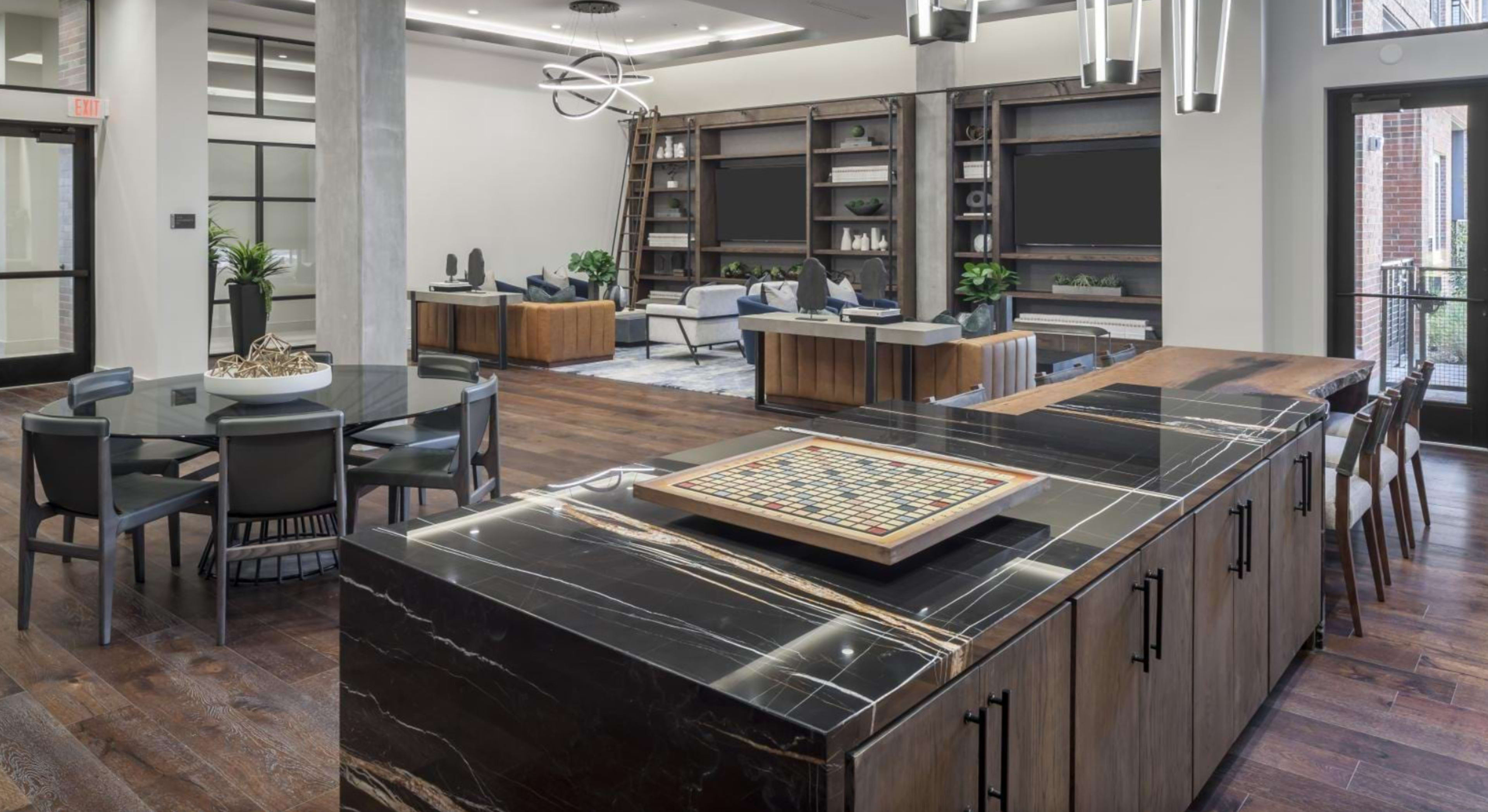 The image shows a modern communal lounge area with a kitchen island, dining table, and seating arrangement, featuring wooden floors and contemporary furnishings.