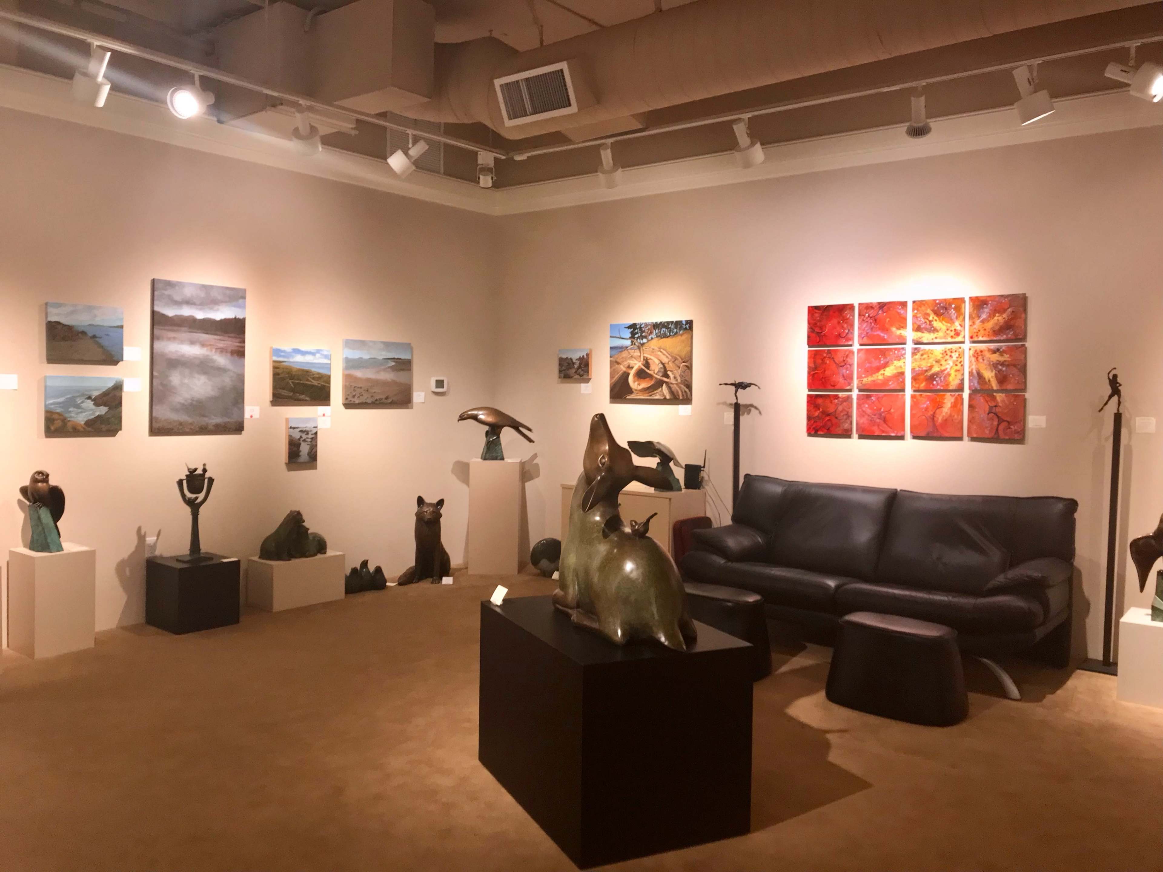 An art gallery featuring various sculptures and paintings displayed on the walls, with a black leather sofa and a round stool in the center.