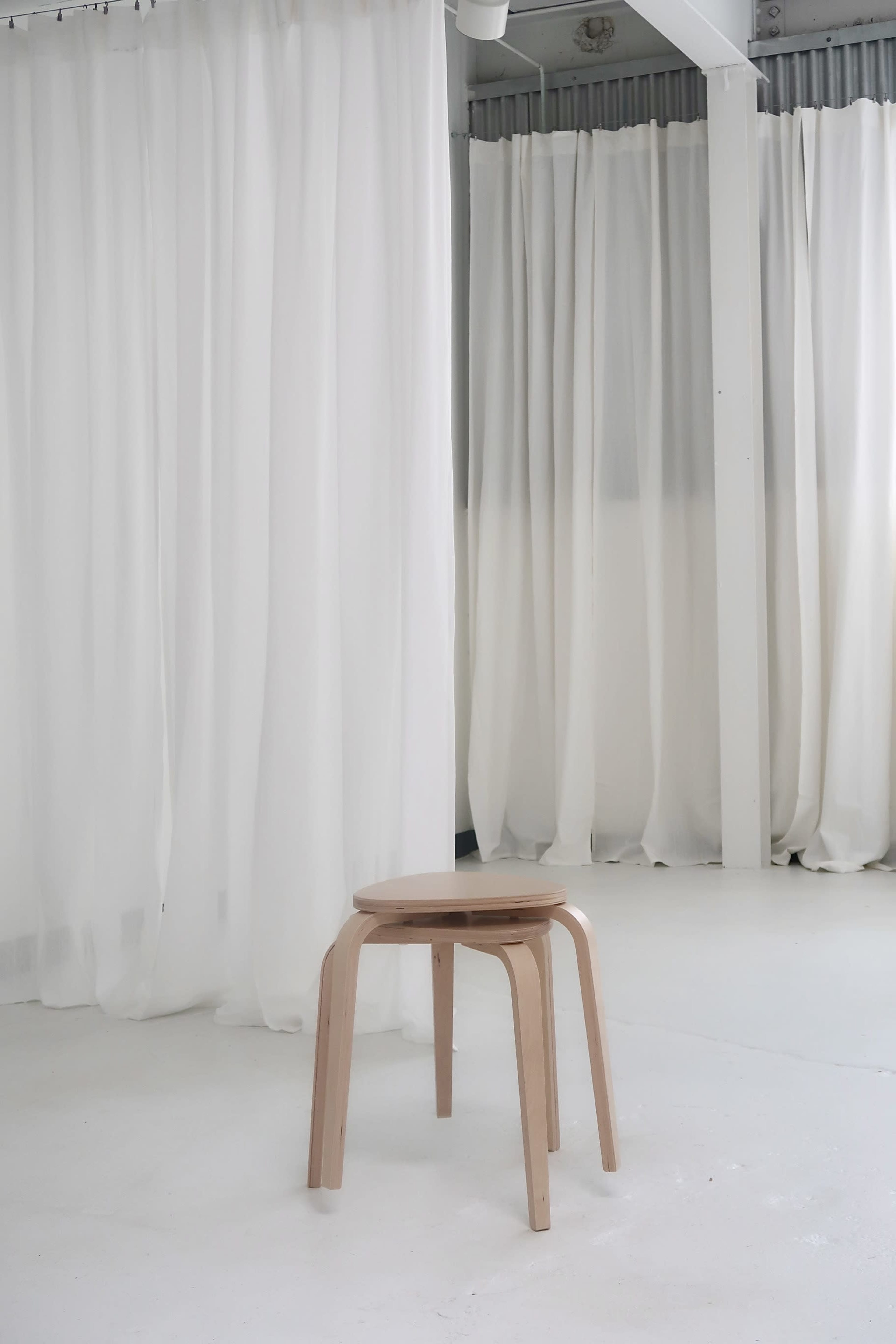 A wooden stool stands on a white floor in front of sheer white curtains.