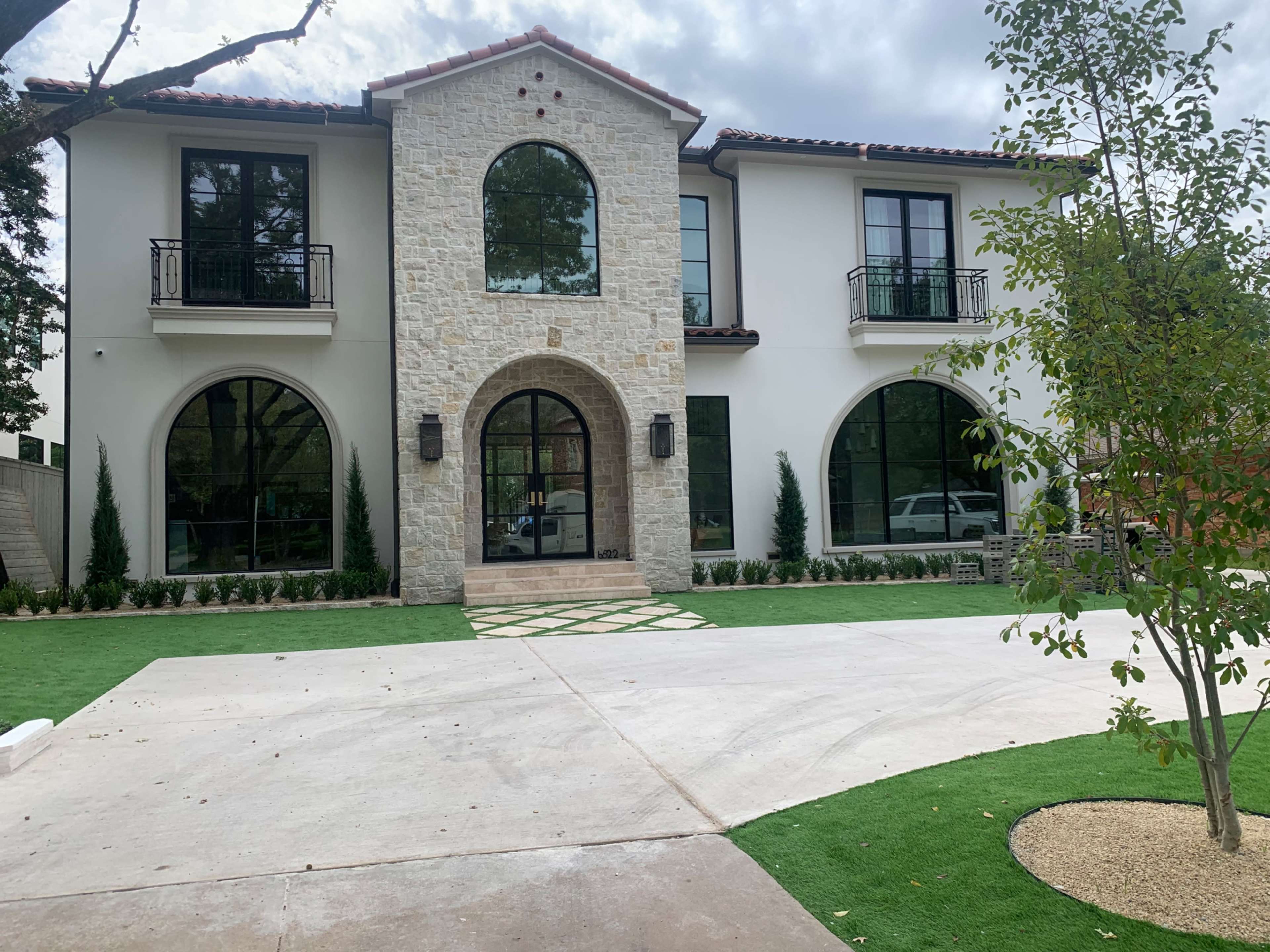 The image shows a modern two-story house with a stone facade, arched windows, and a landscaped front yard.