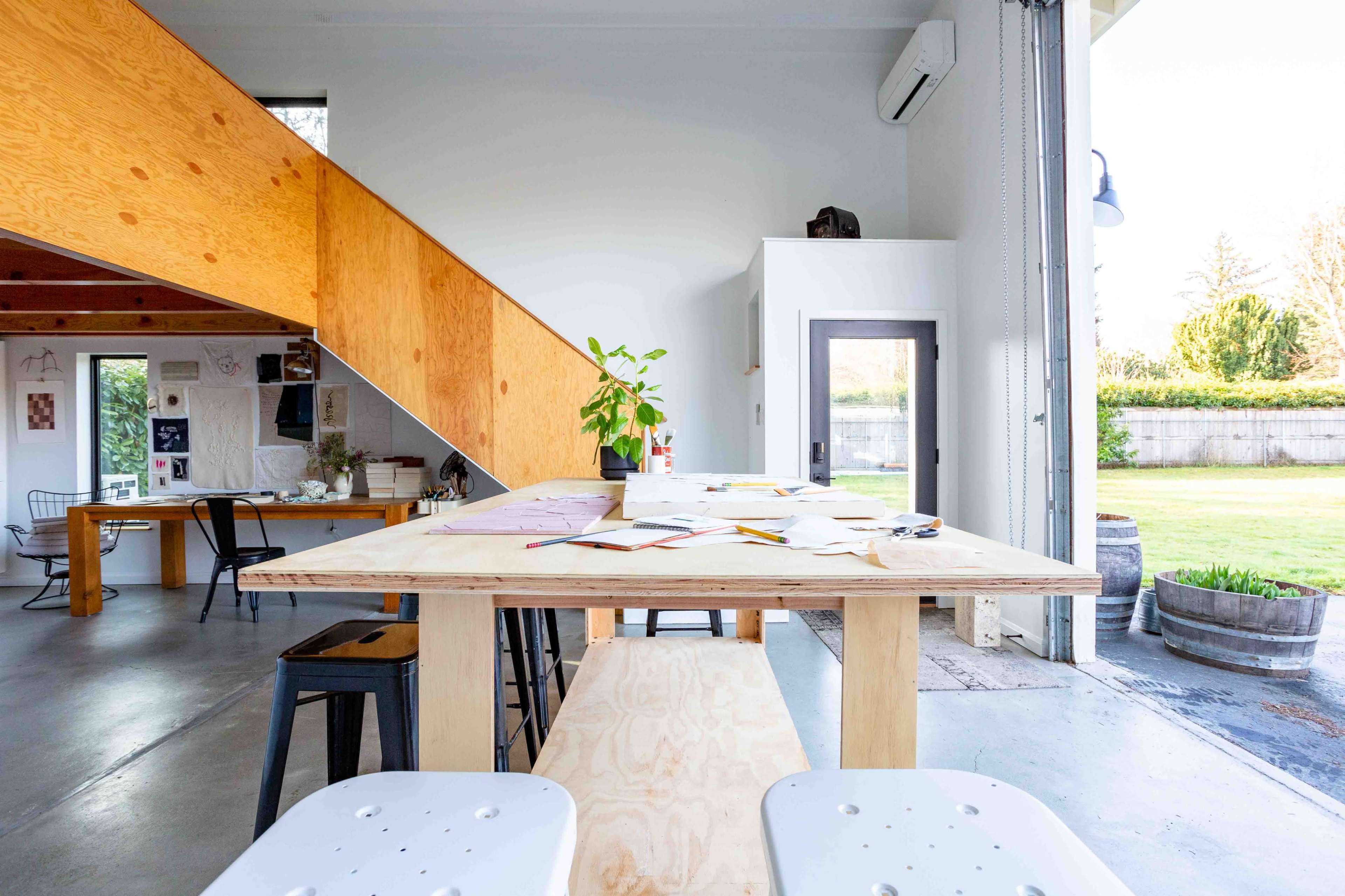 A bright, modern workspace featuring a wooden table with scattered papers, a potted plant, and an open garage door leading to a grassy area.