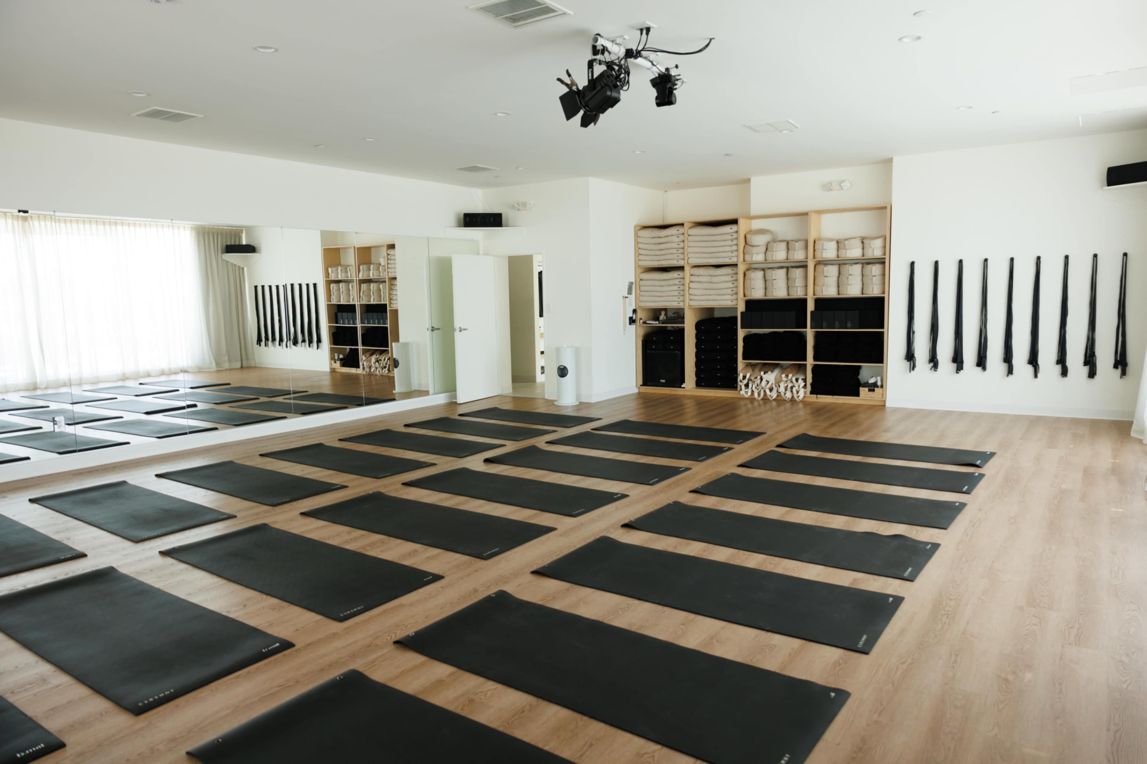 A well-lit yoga studio features rows of black mats arranged on a wooden floor, with mirrors and neatly organized equipment along the walls.