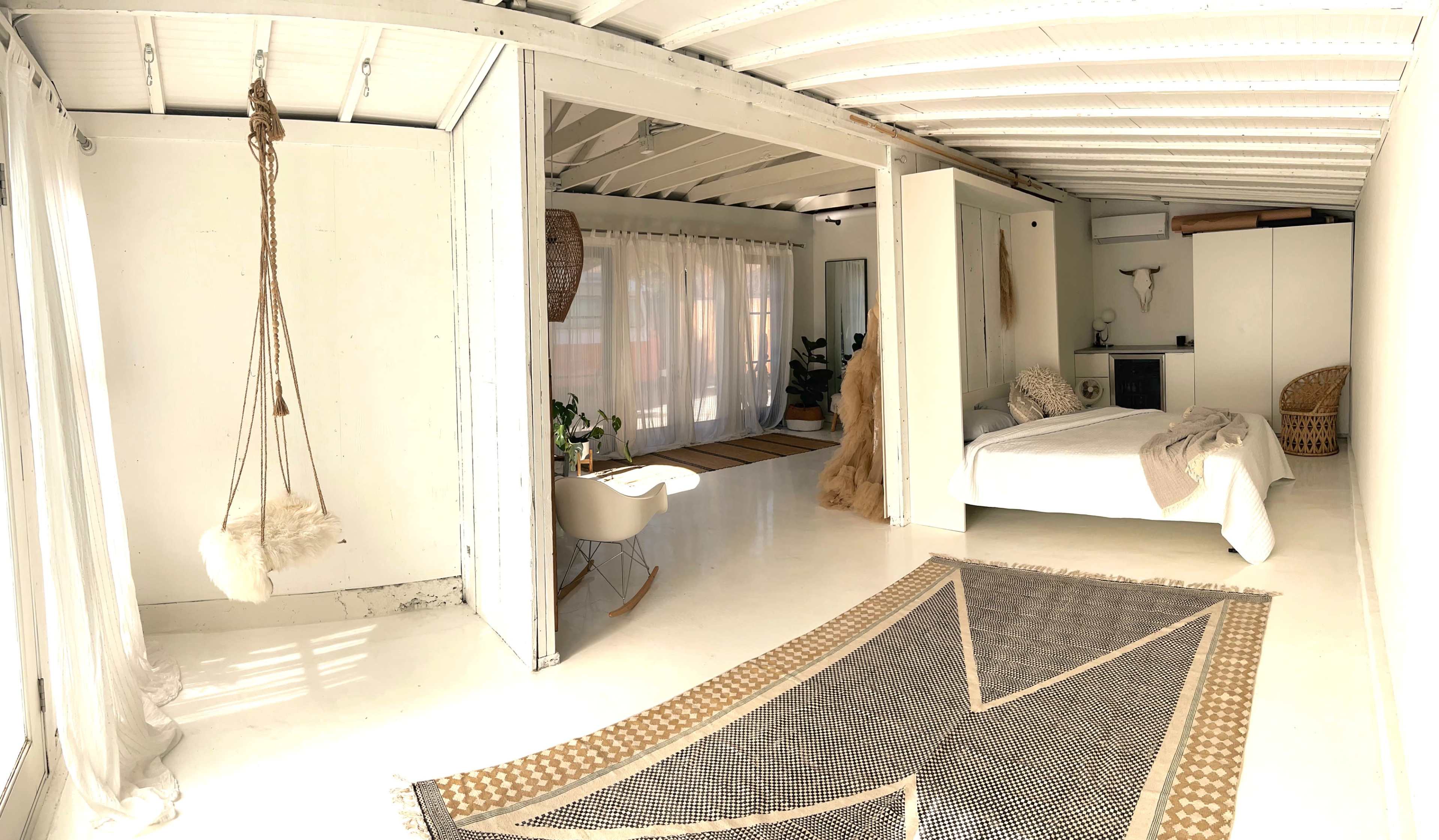 The image shows a modern, minimalistic bedroom with a large bed, a hanging chair, and a woven rug, all within a bright, open space.