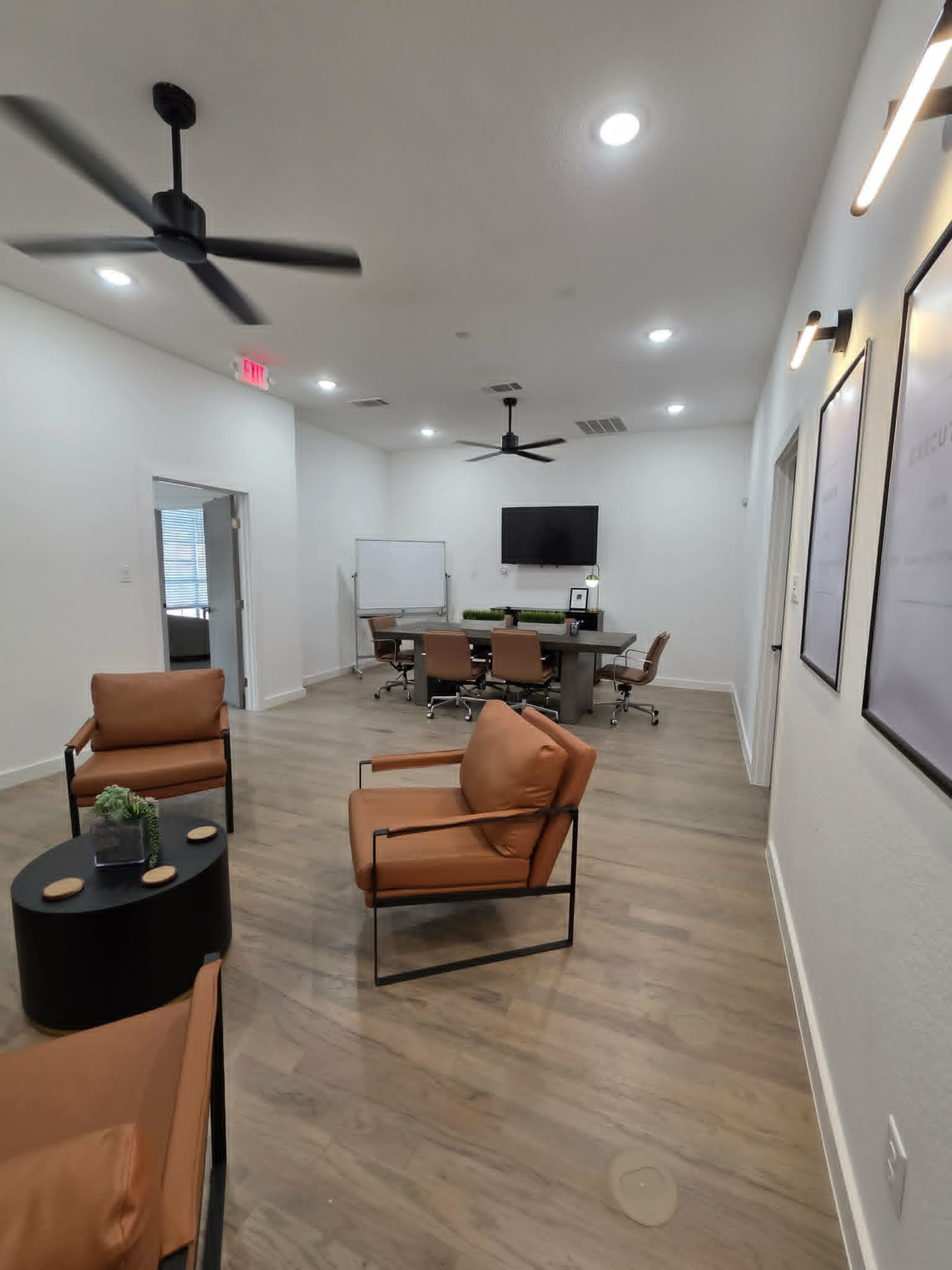 The image shows a modern office space featuring a conference table with chairs, a whiteboard, and several brown lounge chairs arranged around a small black table.