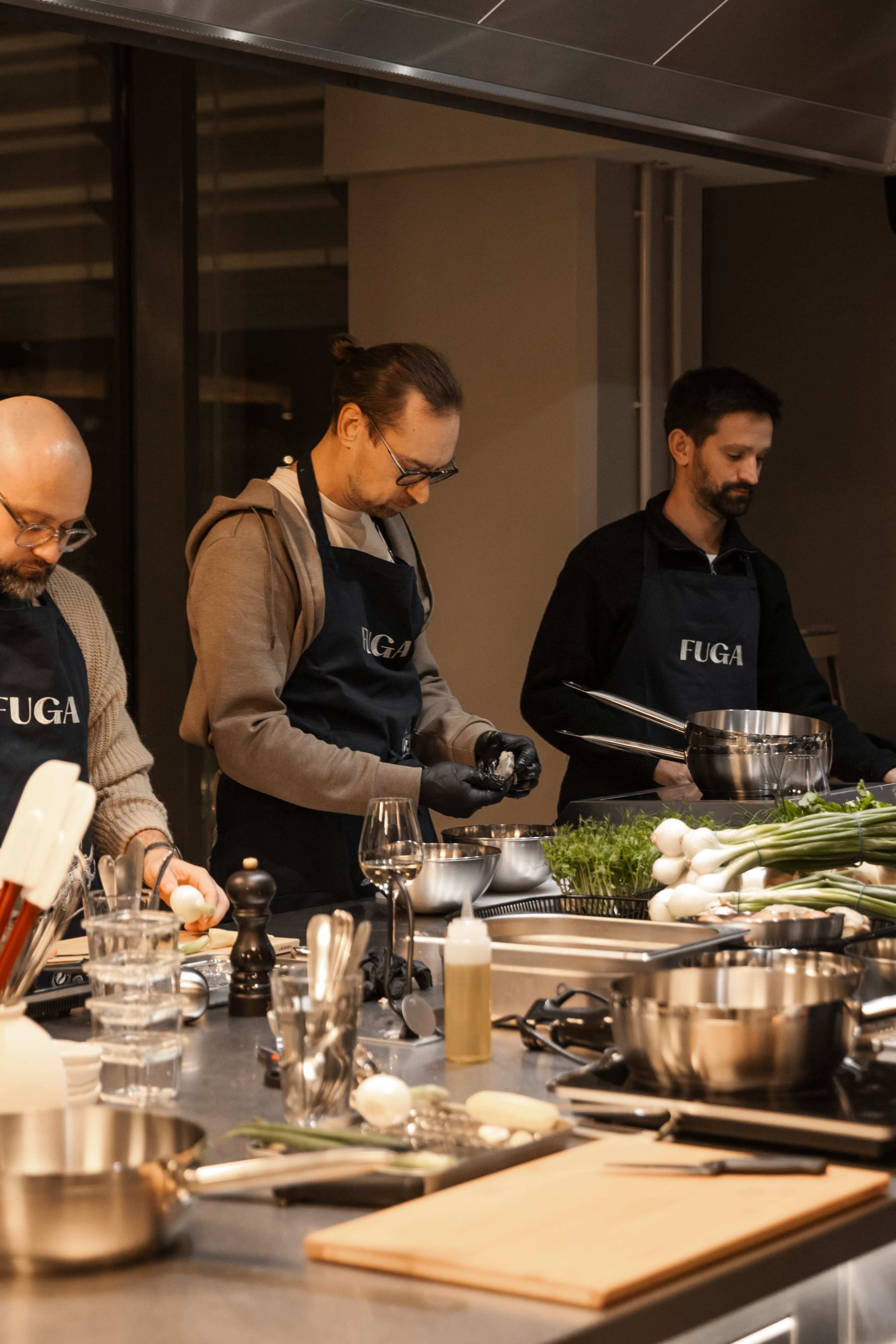 Three men in FUGA aprons prepare food in a modern kitchen equipped with various cooking tools and ingredients.