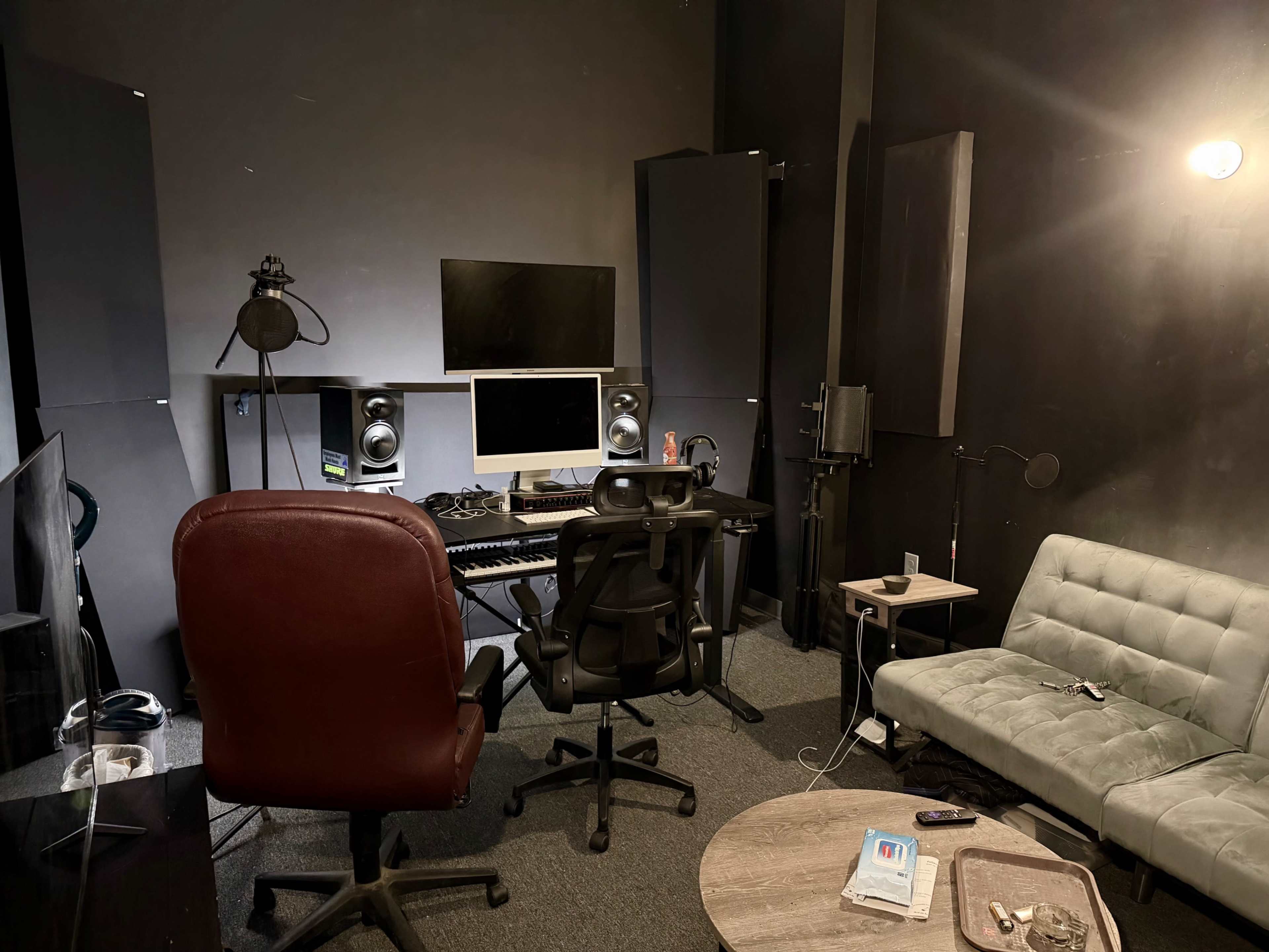 The image shows a small music production studio with a computer setup, audio monitors, a microphone, and seating arrangements.