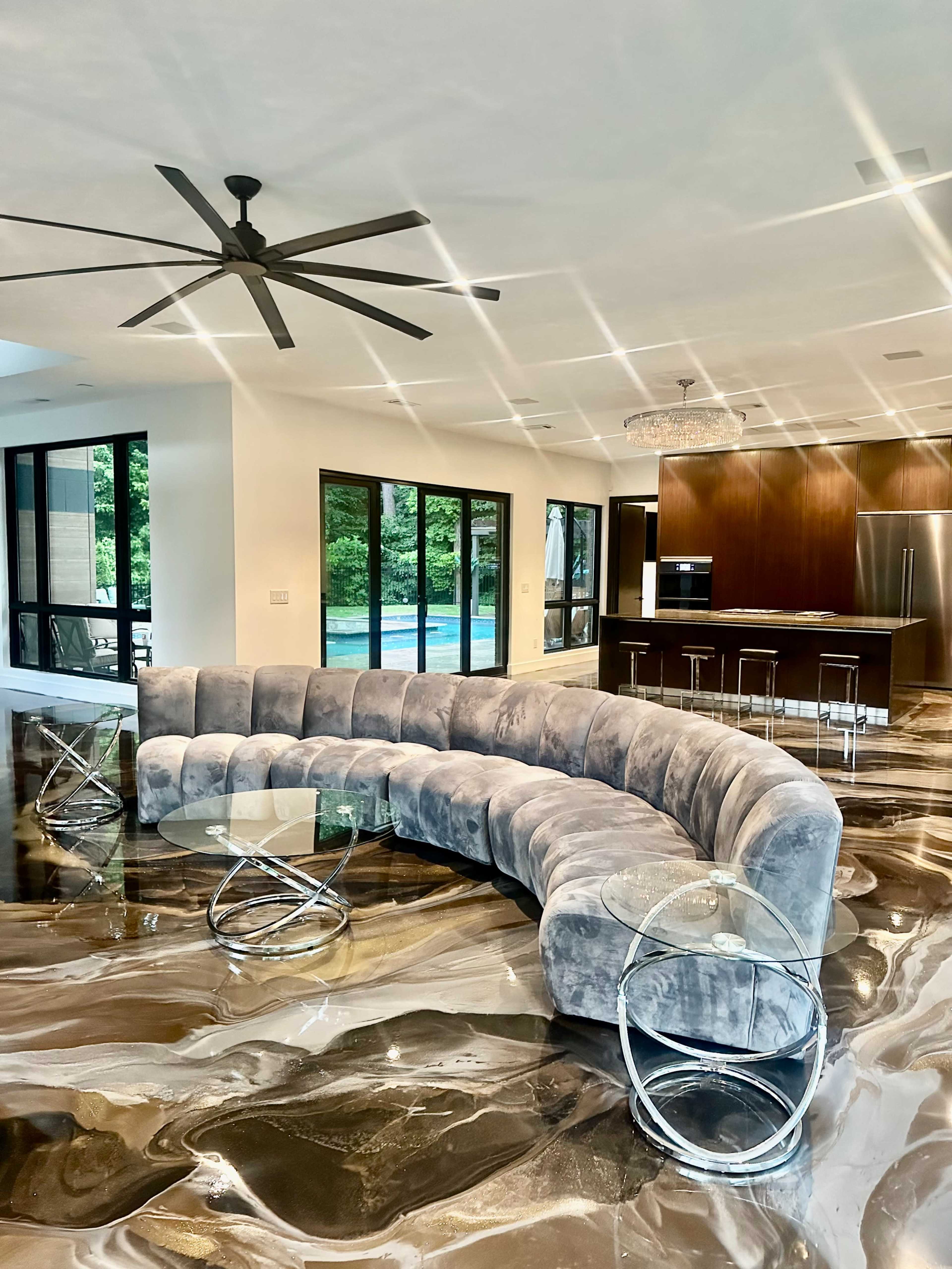 A curved gray sofa and clear glass tables are situated in a modern living space with shiny marble flooring, large windows, and a view of an outdoor pool.